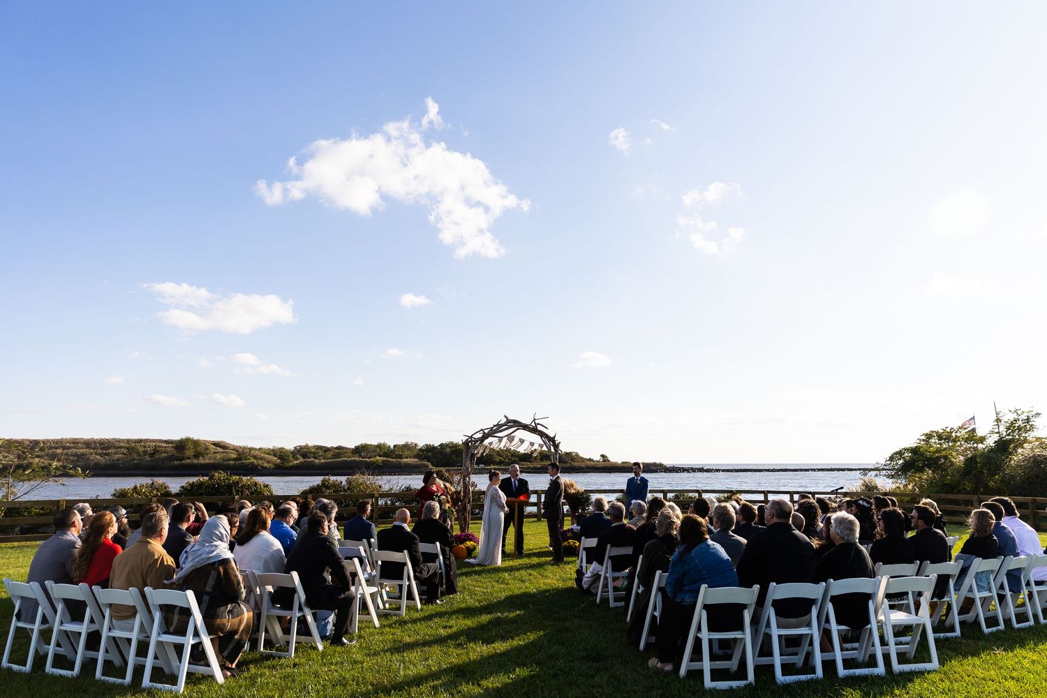 Wide shot of outdoor wedding ceremony with white chairs and guests facing rustic arch by waterfront.