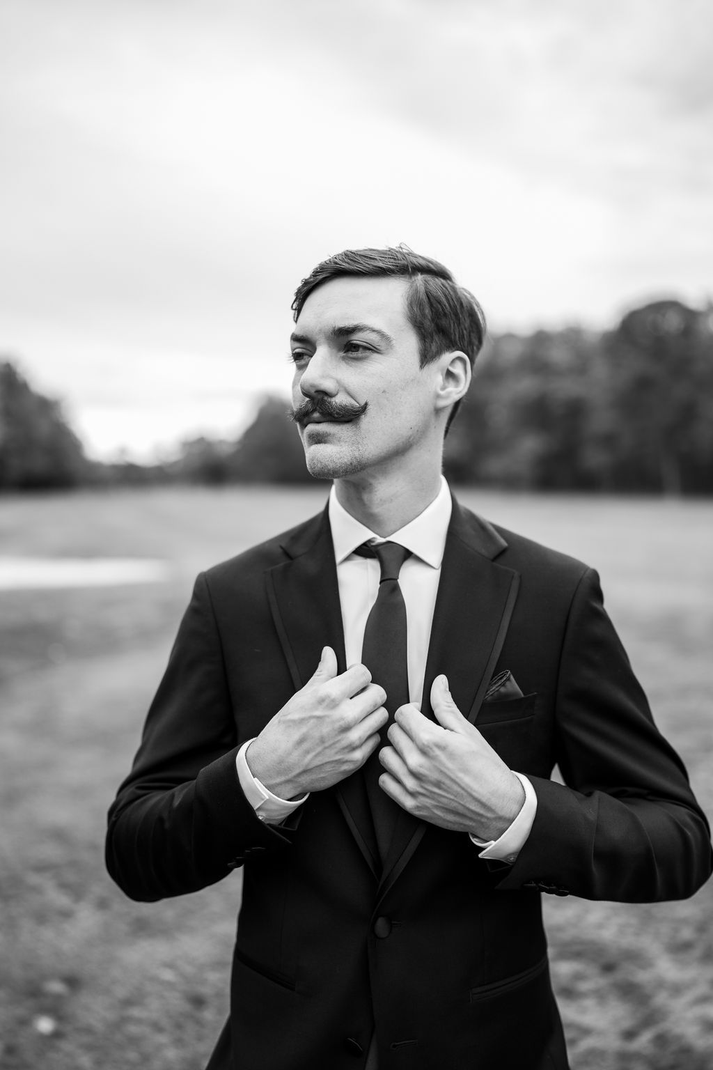 Dapper groom with mustache adjusts suit jacket in black and white outdoor portrait.