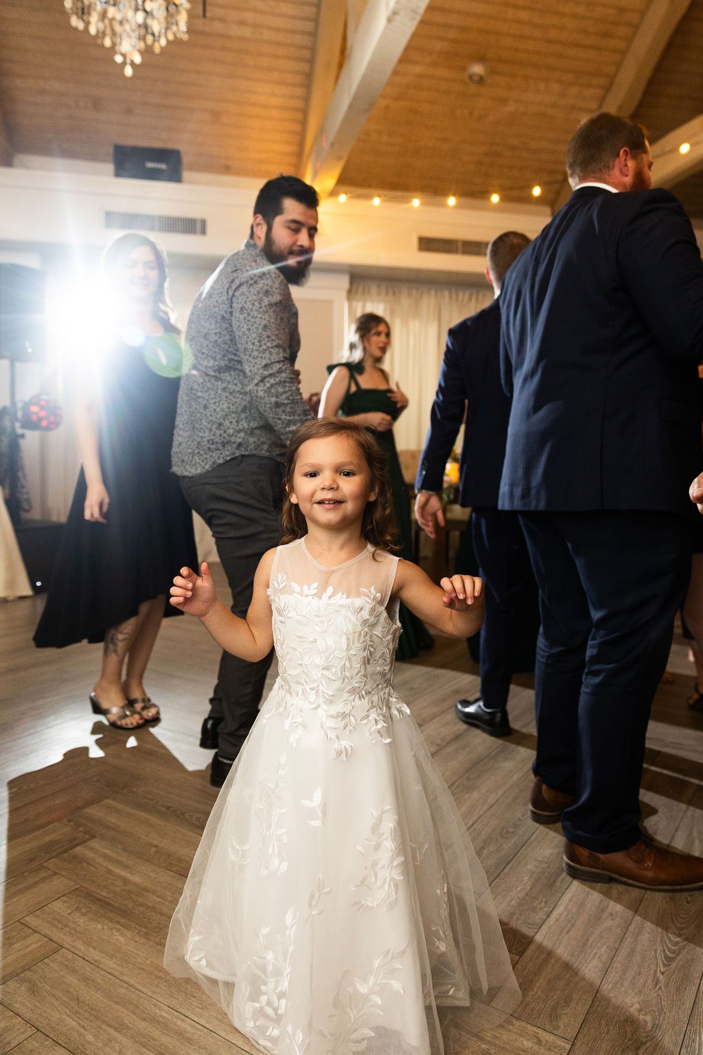 Adorable flower girl in a white lace dress smiles while dancing among guests at a wedding reception.