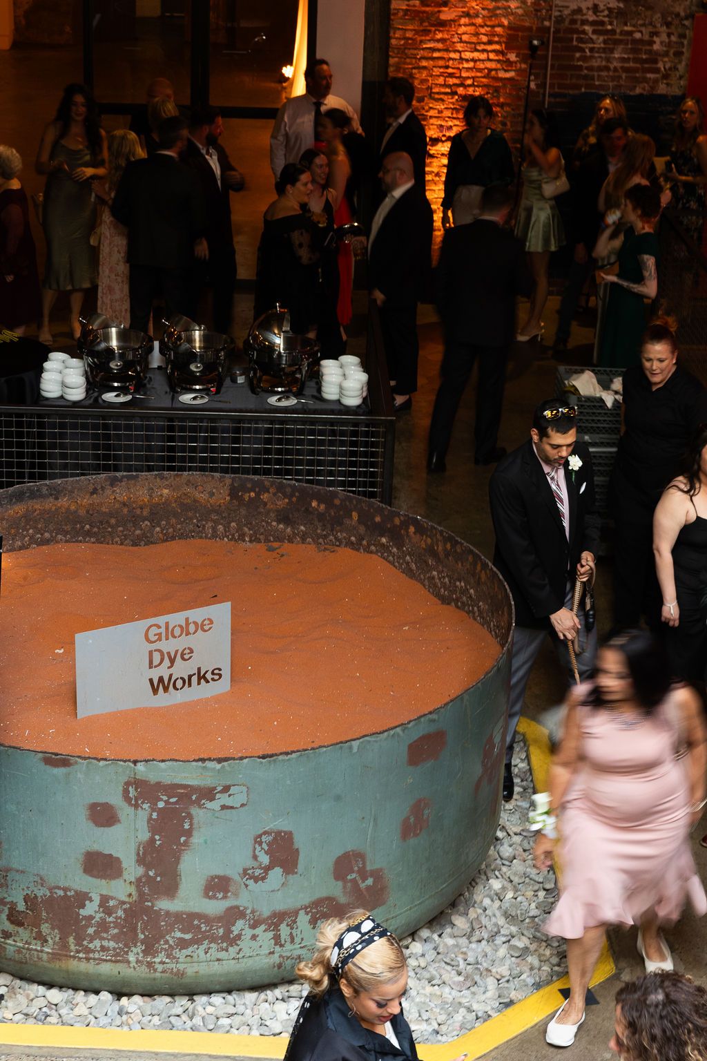 Wedding guests socializing around a large rustic vat at Globe Dye Works venue.
