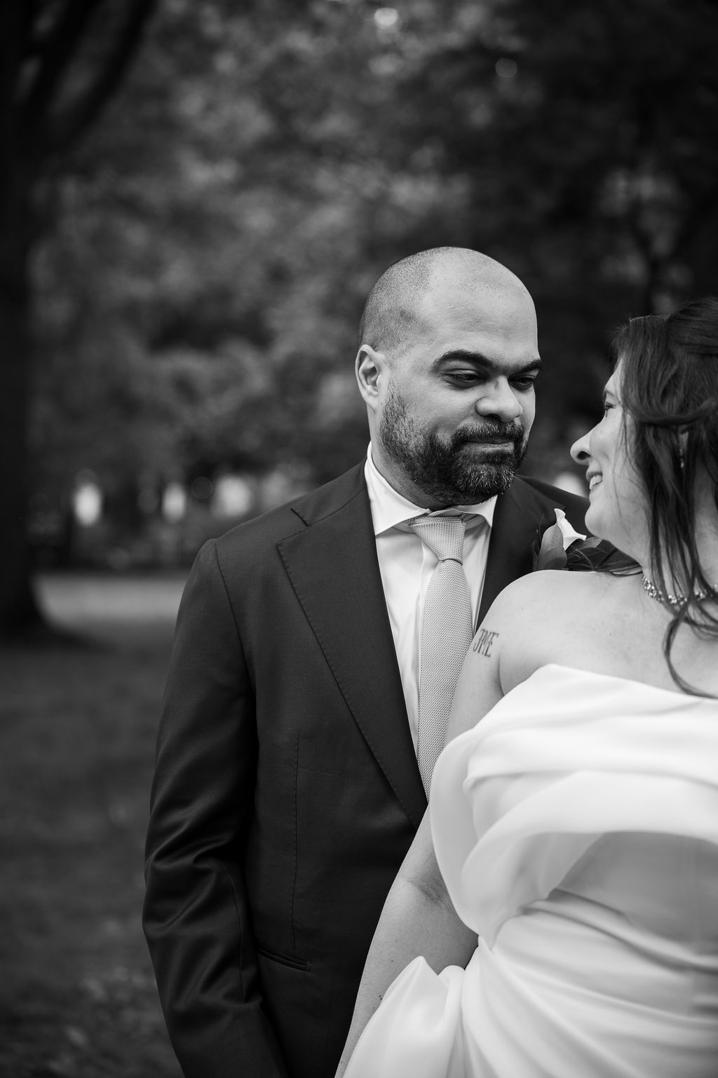 Black and white photo of a groom in dark suit gazing at his smiling bride in white wedding dress outdoors.