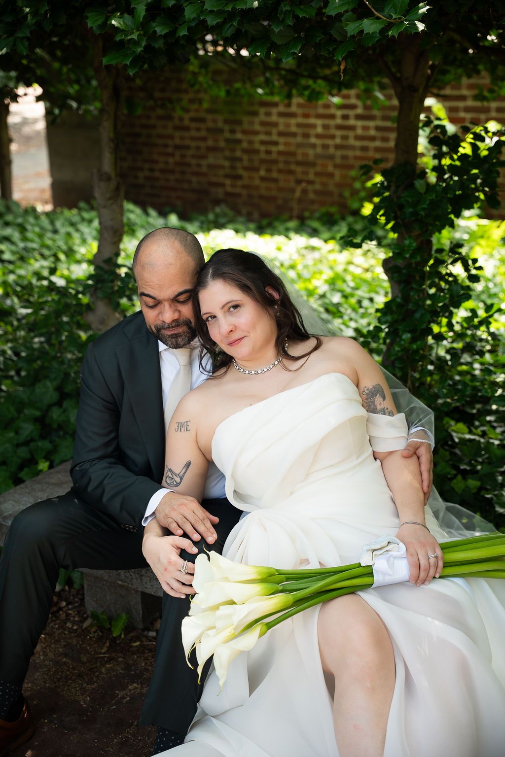 Bride and groom sitting on bench in garden, bride holding white calla lily bouquet, groom embracing her tenderly.
