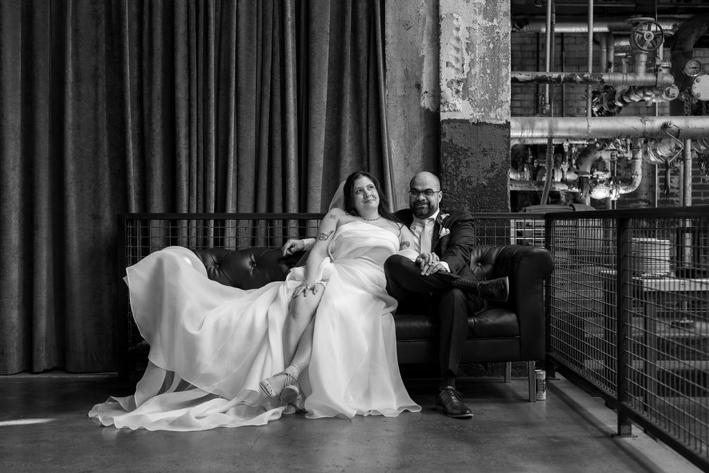 Bride and groom relax on leather sofa in black and white portrait at industrial wedding venue.