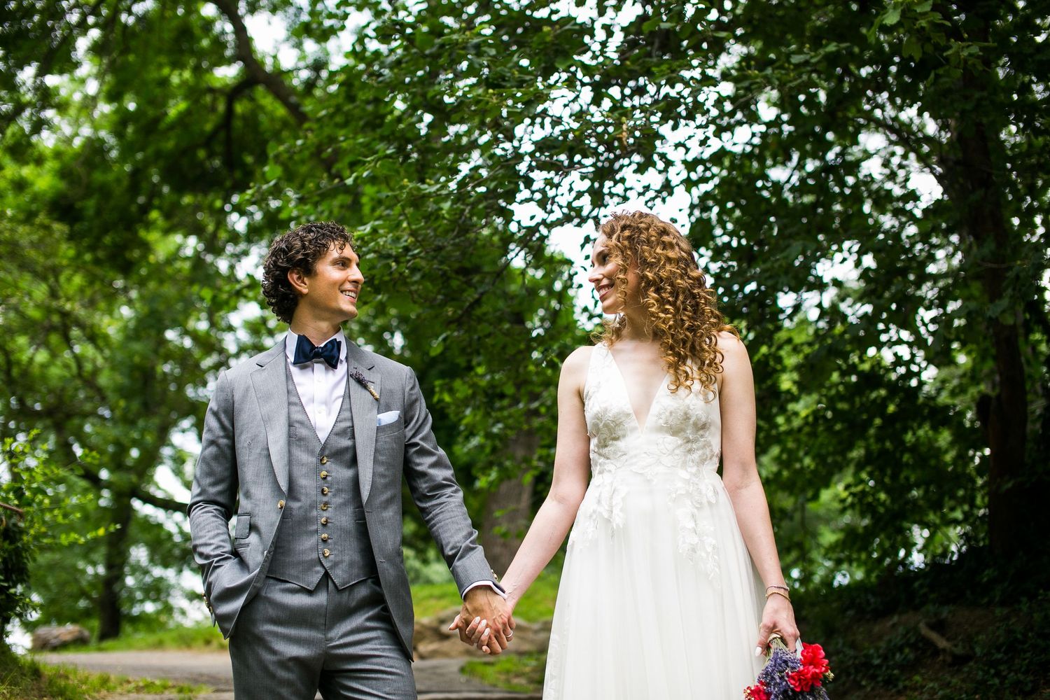Smiling newlyweds strolling along a tree-lined path, bride in elegant white dress, groom in gray suit.