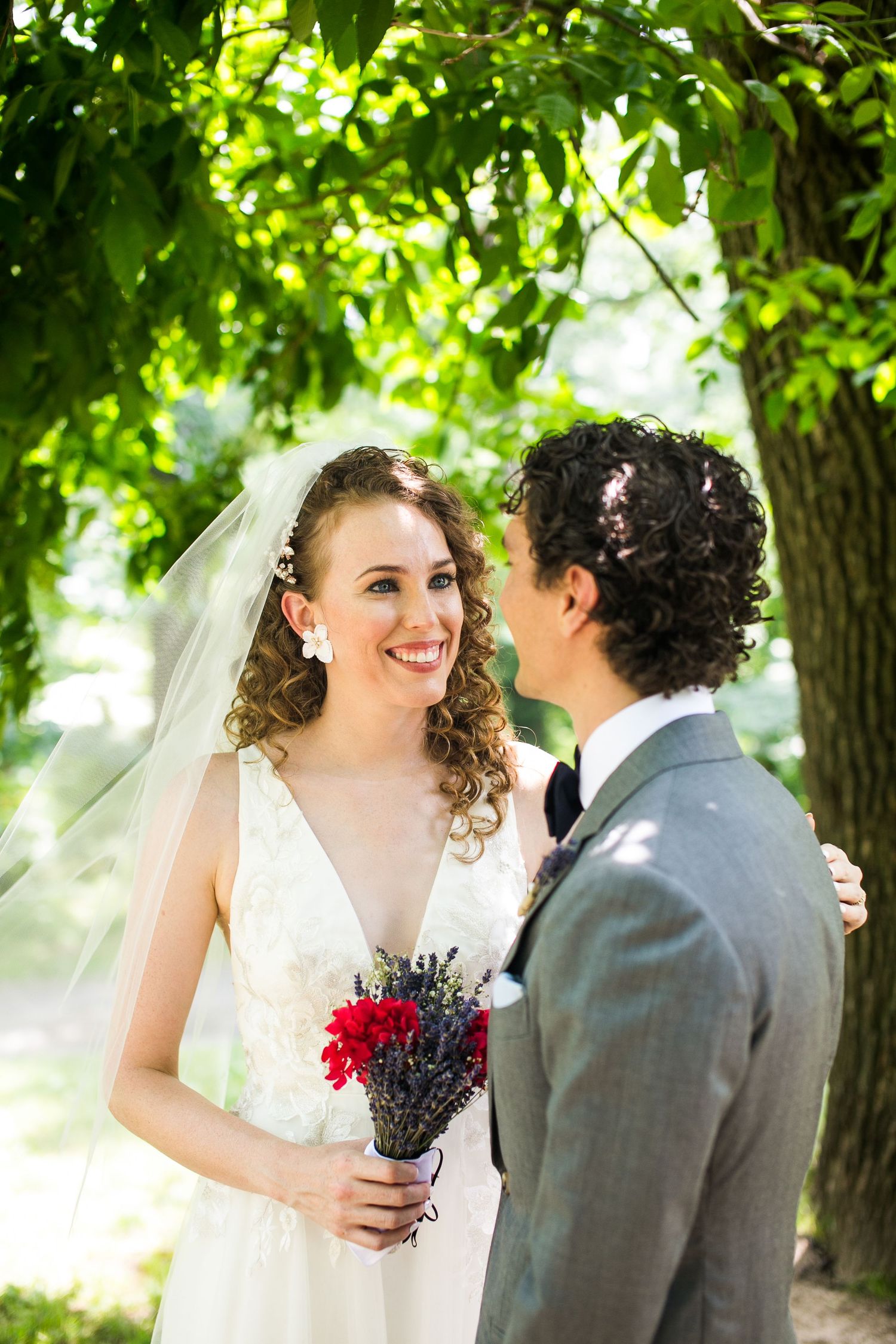 Smiling bride in white gown holding red and lavender bouquet gazes at groom in gray suit outdoors.