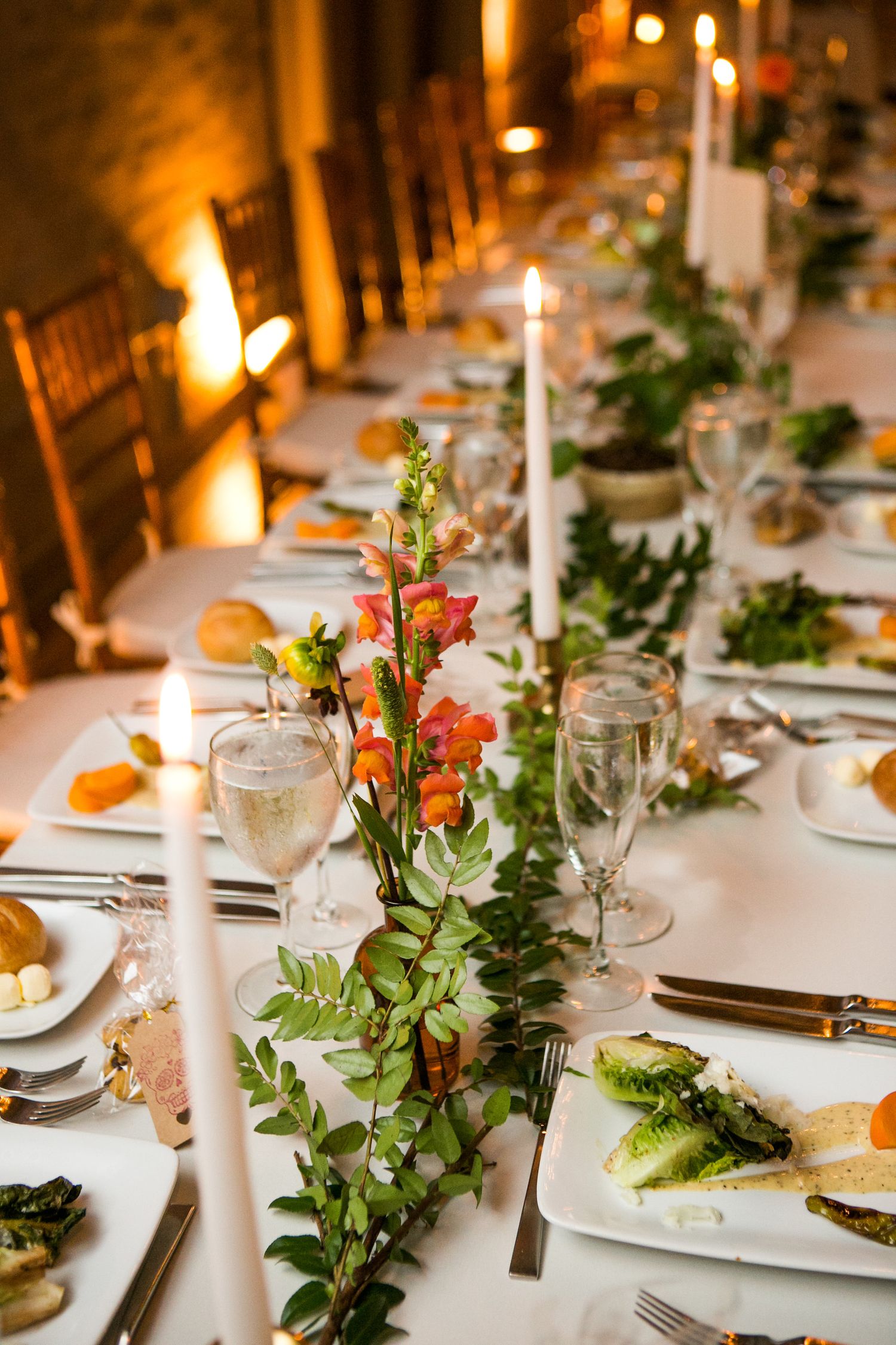 Long wedding reception table with candles, greenery, and orange floral centerpieces in warm lighting.