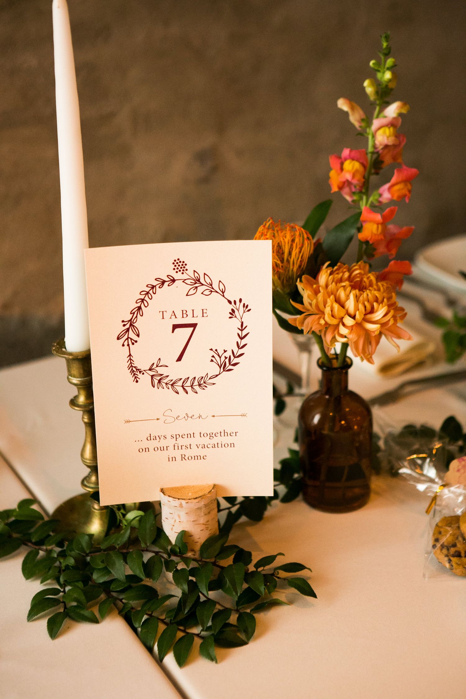 Wedding table number seven card with wreath design beside amber bottle florals and brass candlestick centerpiece.