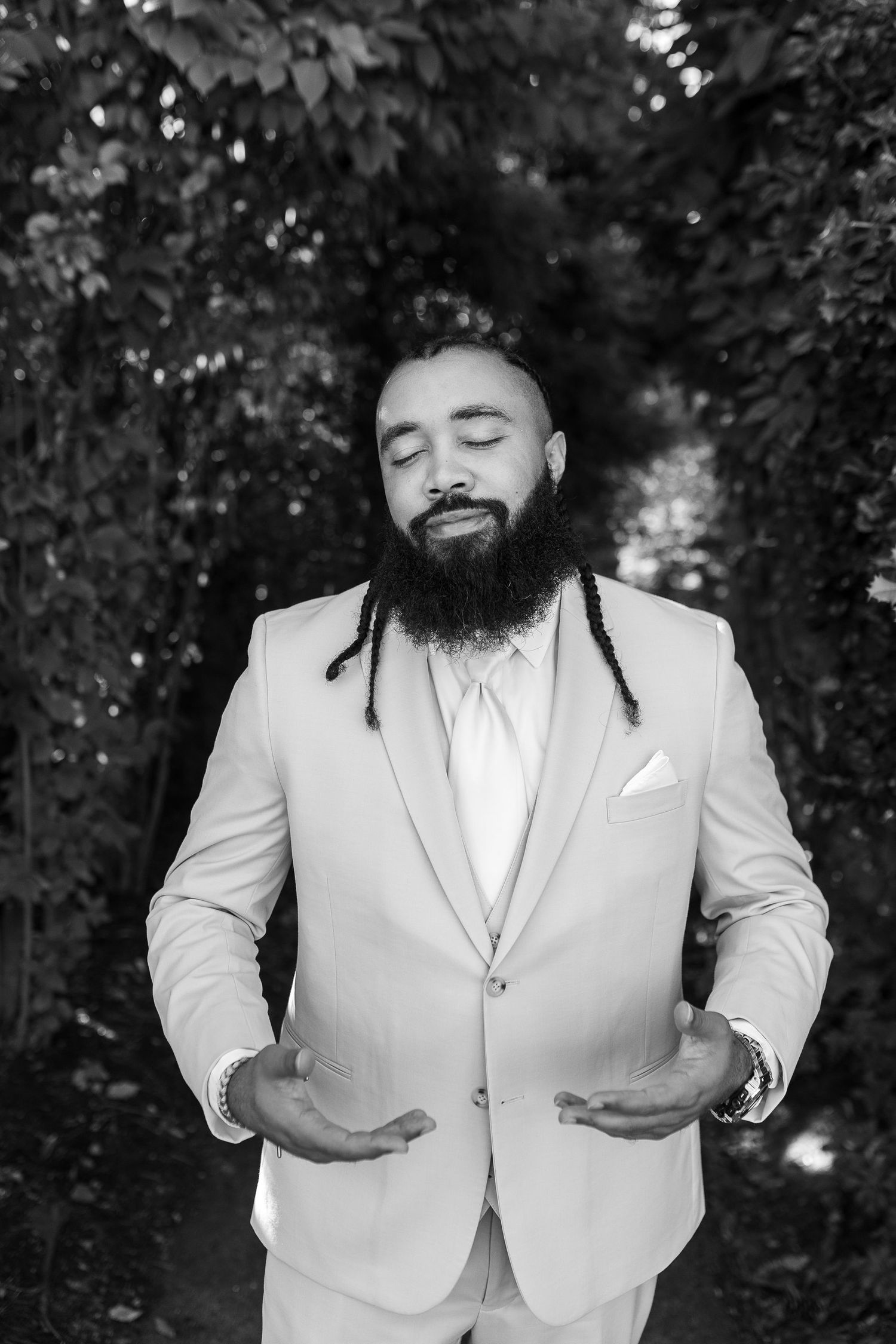 Black and white portrait of bearded groom in light suit with eyes closed outdoors.