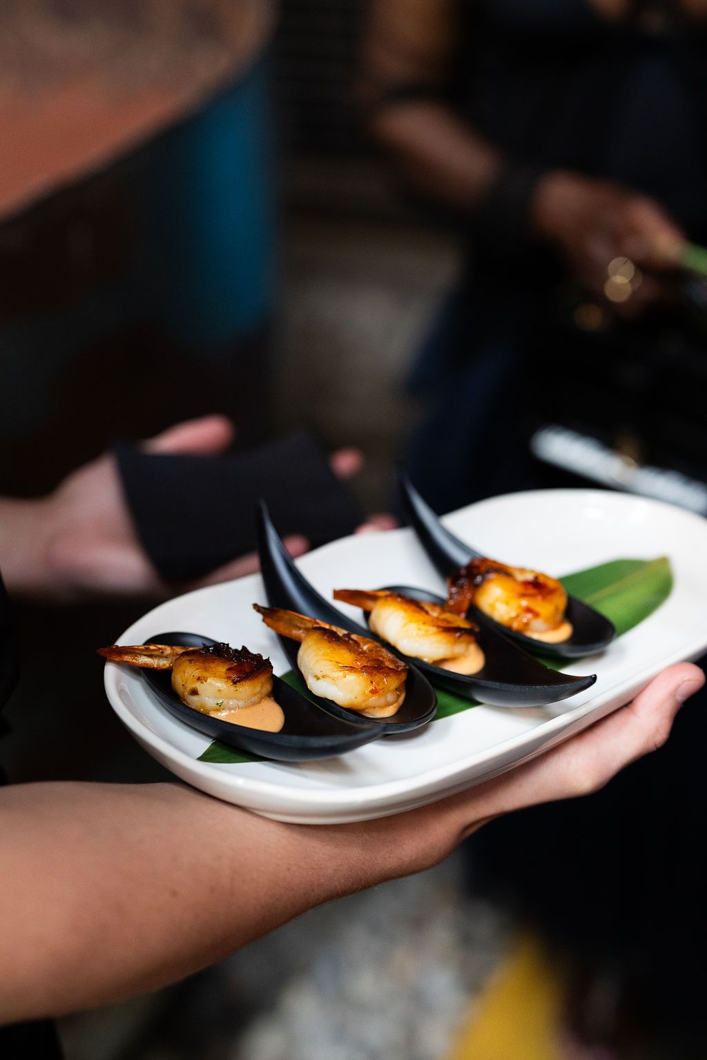 Server presenting elegant appetizers of glazed scallops in black shells on a white plate.