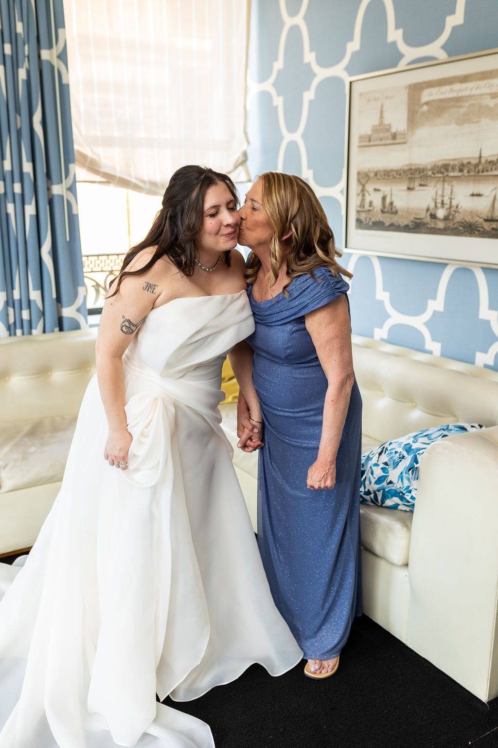 Bride in white strapless gown receiving a kiss from her mother in a blue and white decorated room.