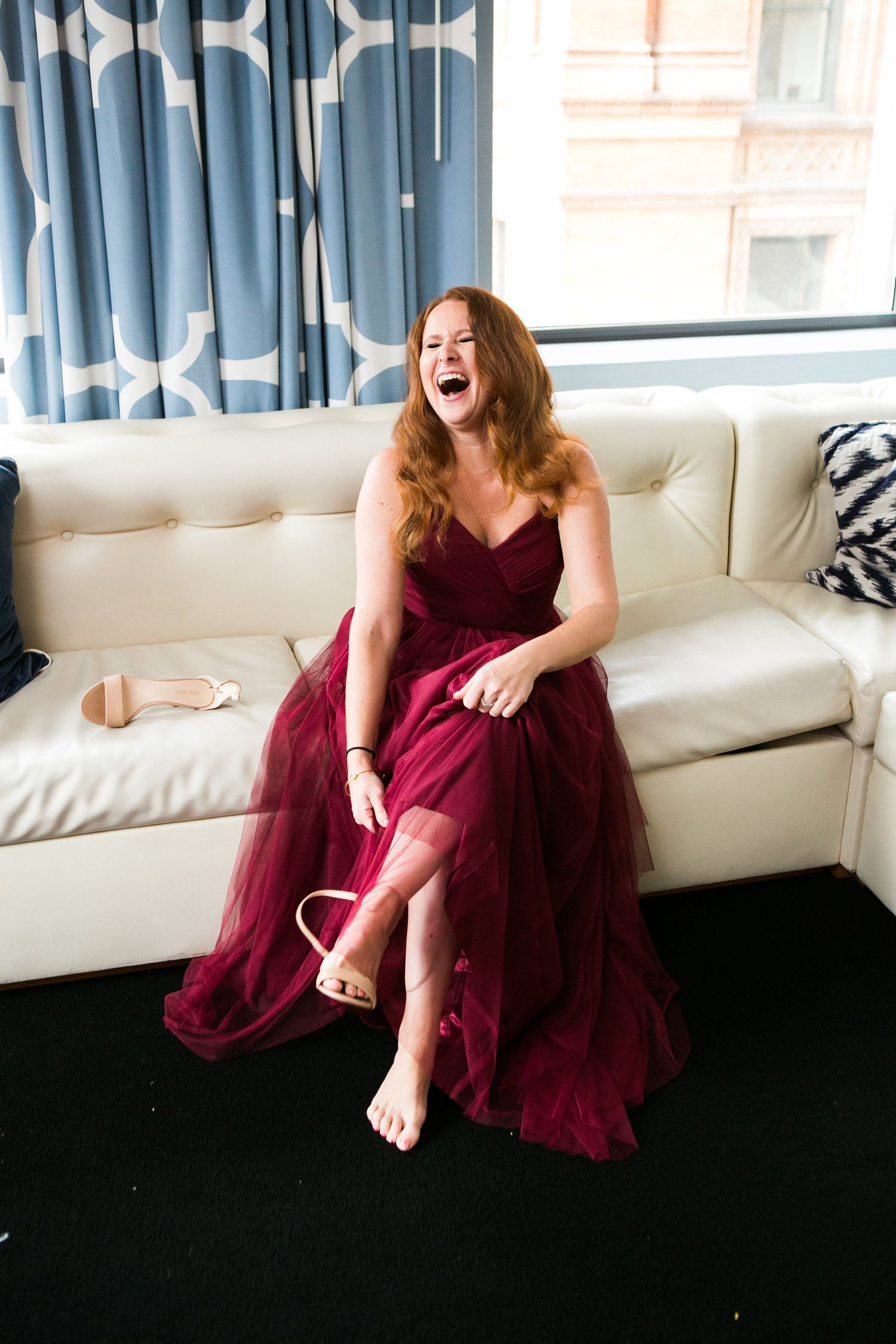 Laughing bridesmaid in a burgundy tulle gown sits on a white leather sofa while putting on her heels.