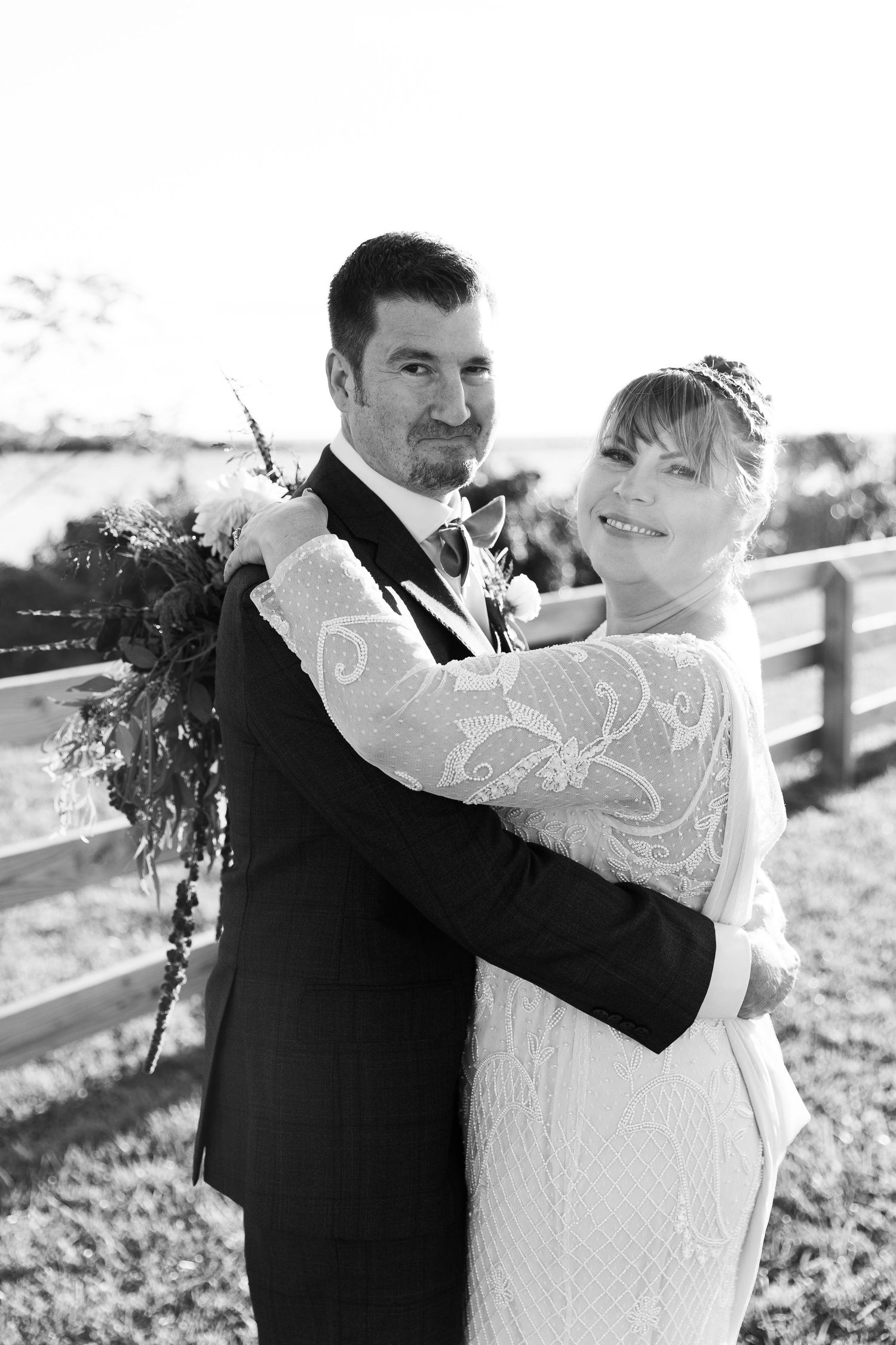 Black and white photo of bride and groom embracing outdoors at wedding ceremony near wooden fence.