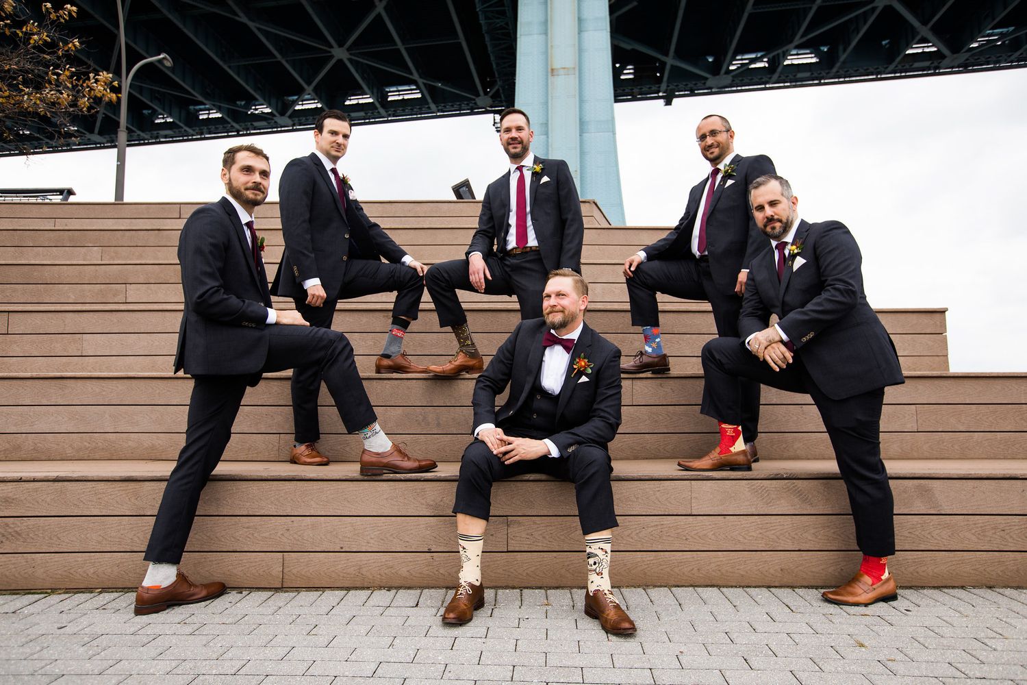 Six groomsmen in black suits pose on outdoor steps showing off colorful socks at a wedding.