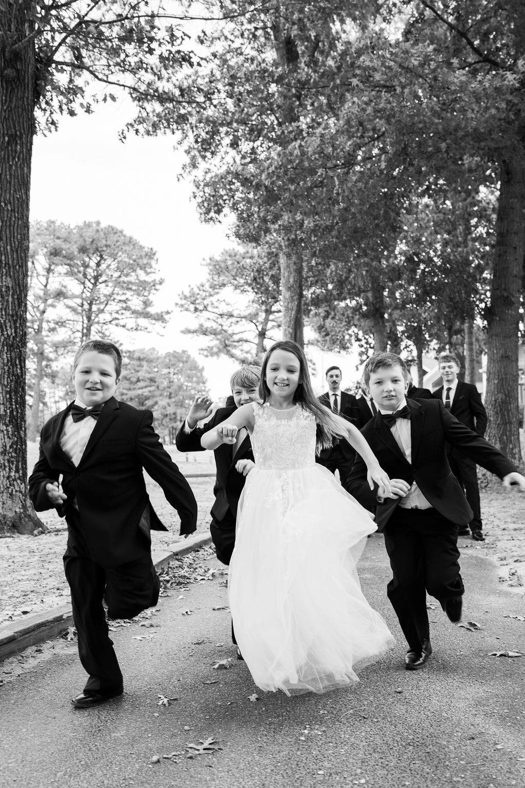 Children in formal wedding attire run joyfully down a tree-lined path in black and white photo.