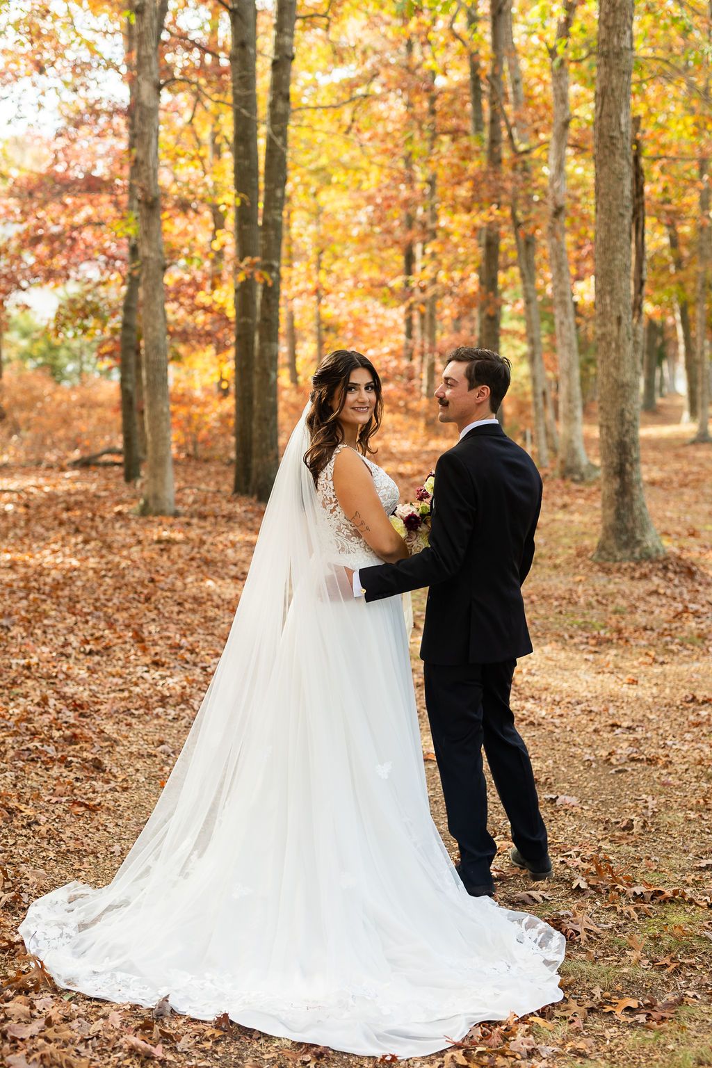 Bride and groom holding hands in autumn forest, bride wearing lace wedding gown with long veil and train.