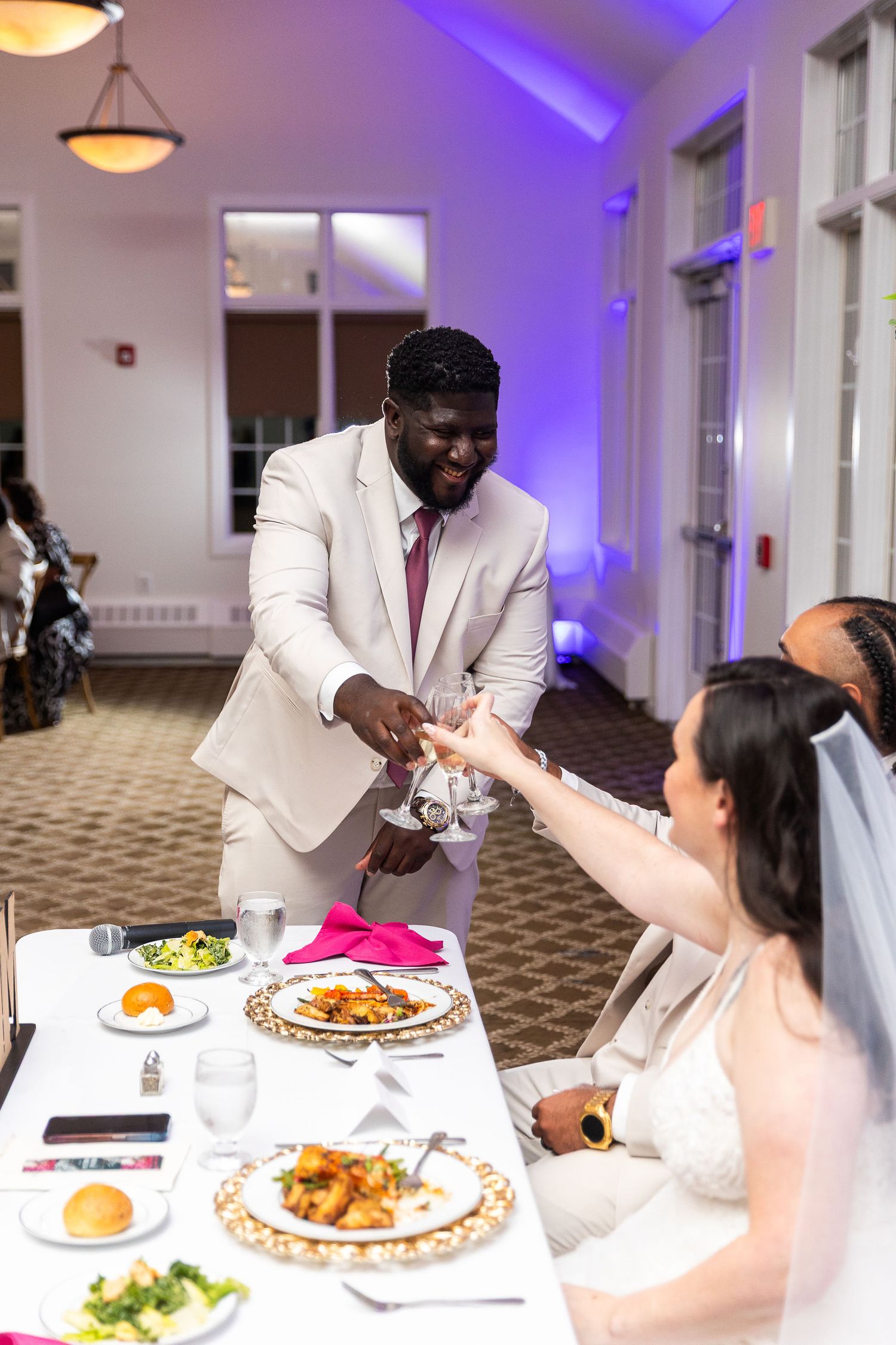 A groom in a beige suit hands a drink to his bride at the wedding reception head table.