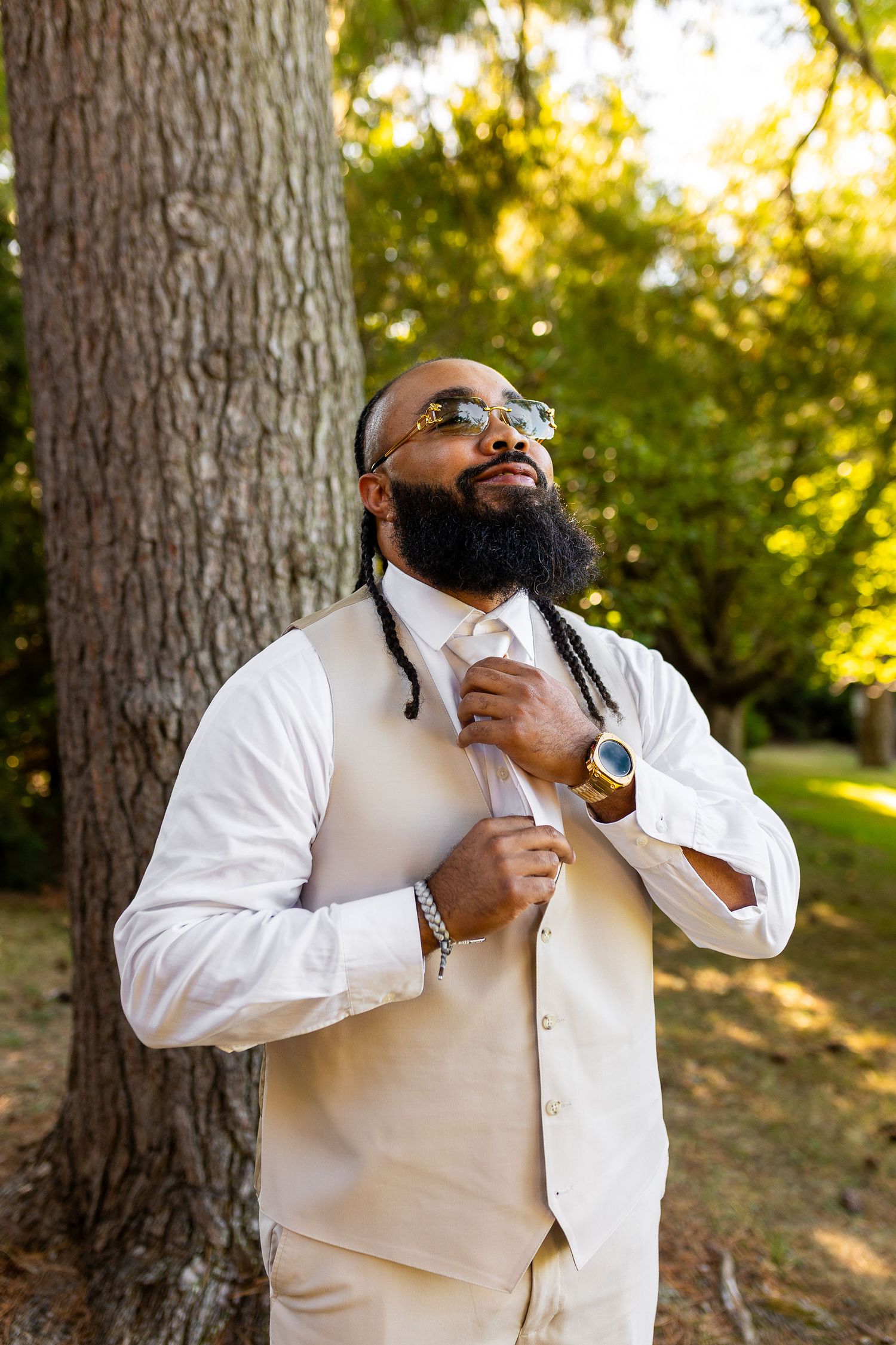 Stylish groom with dreadlocks and beard in cream suit and sunglasses adjusting his tie outdoors.