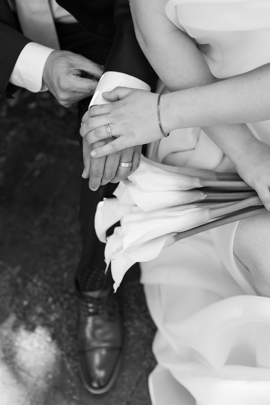 Close-up black and white photo of bride and groom's hands with wedding rings resting on white calla lily bouquet.