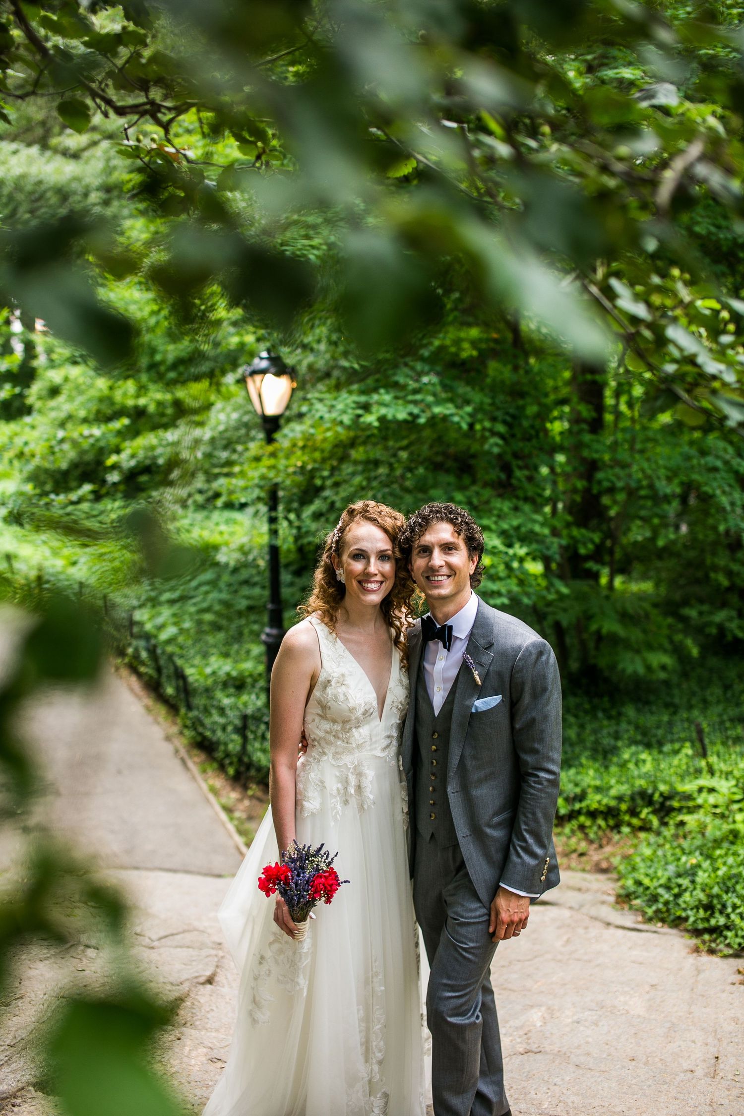 Bride and groom smiling on a lush green park path, bride holding red bouquet, groom in gray suit with bow tie.