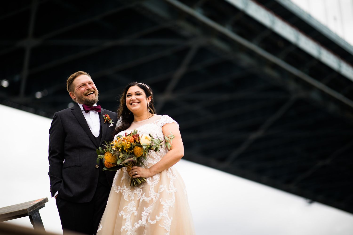 Bride and groom smiling together beneath a large bridge, bride holding a colorful fall bouquet.