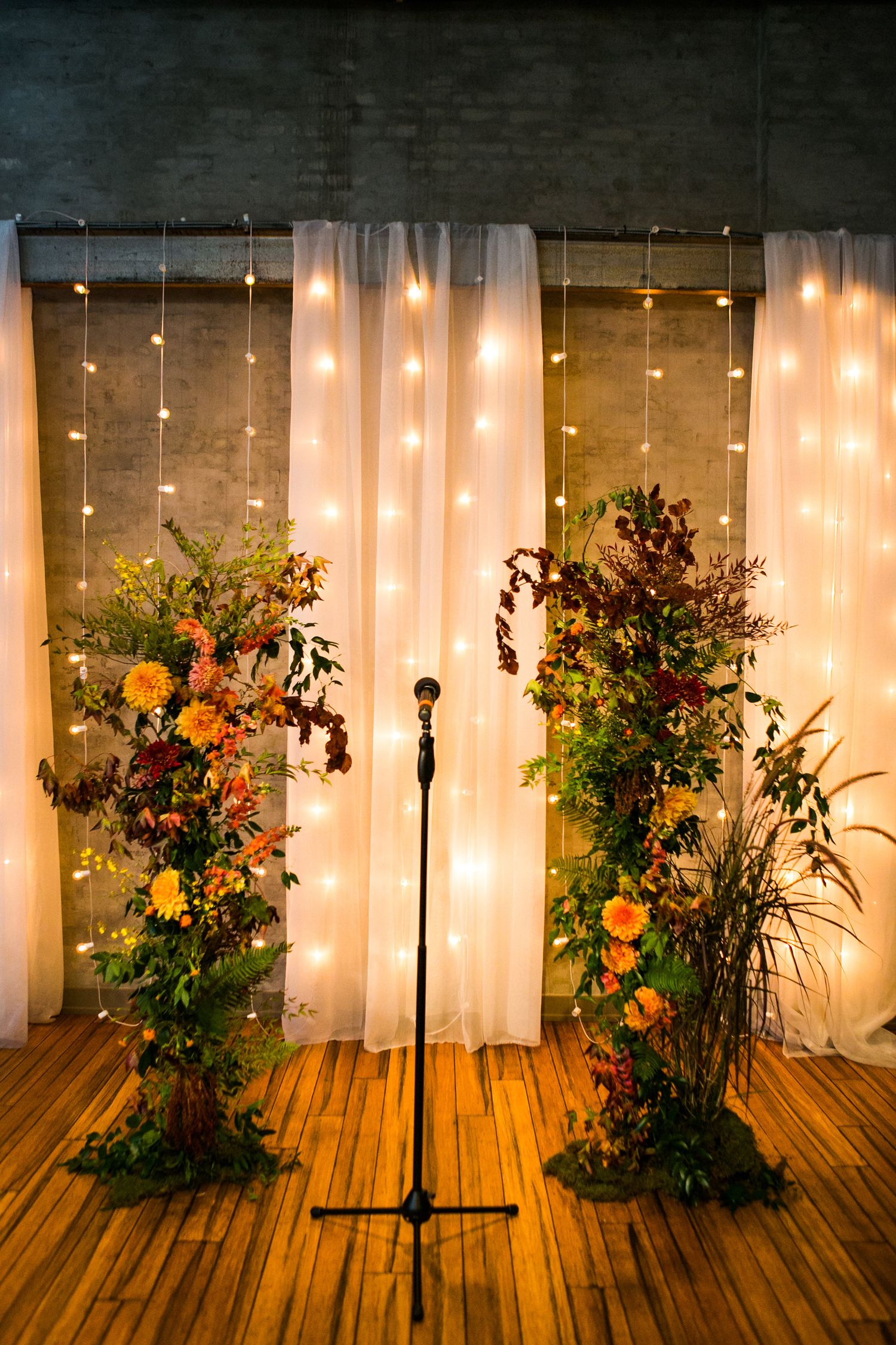 Elegant wedding ceremony backdrop with fairy lights, white draping, and lush autumn floral arrangements on wooden floor.