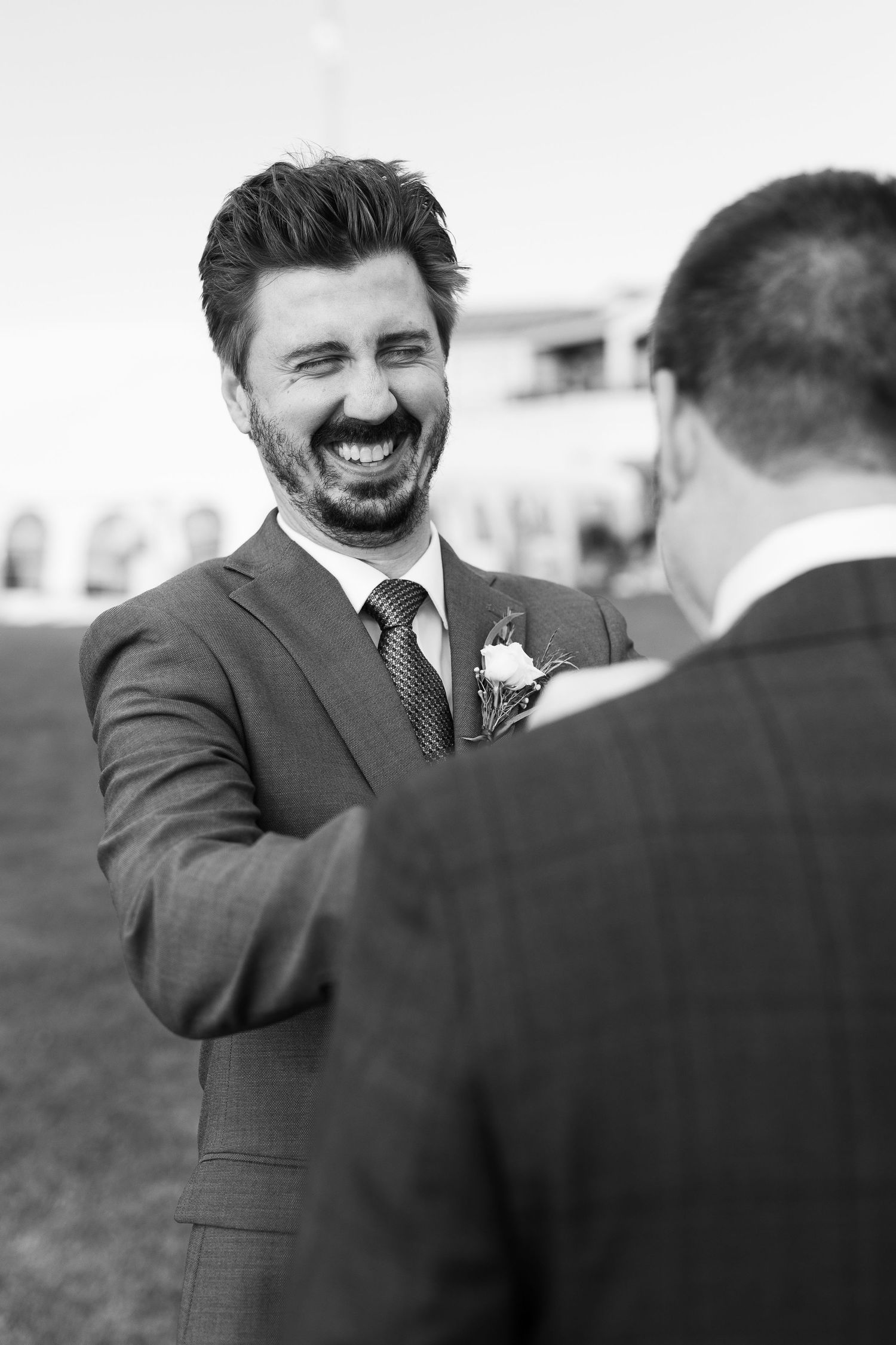 Black and white photo of bearded groom laughing joyfully with guest at outdoor wedding.