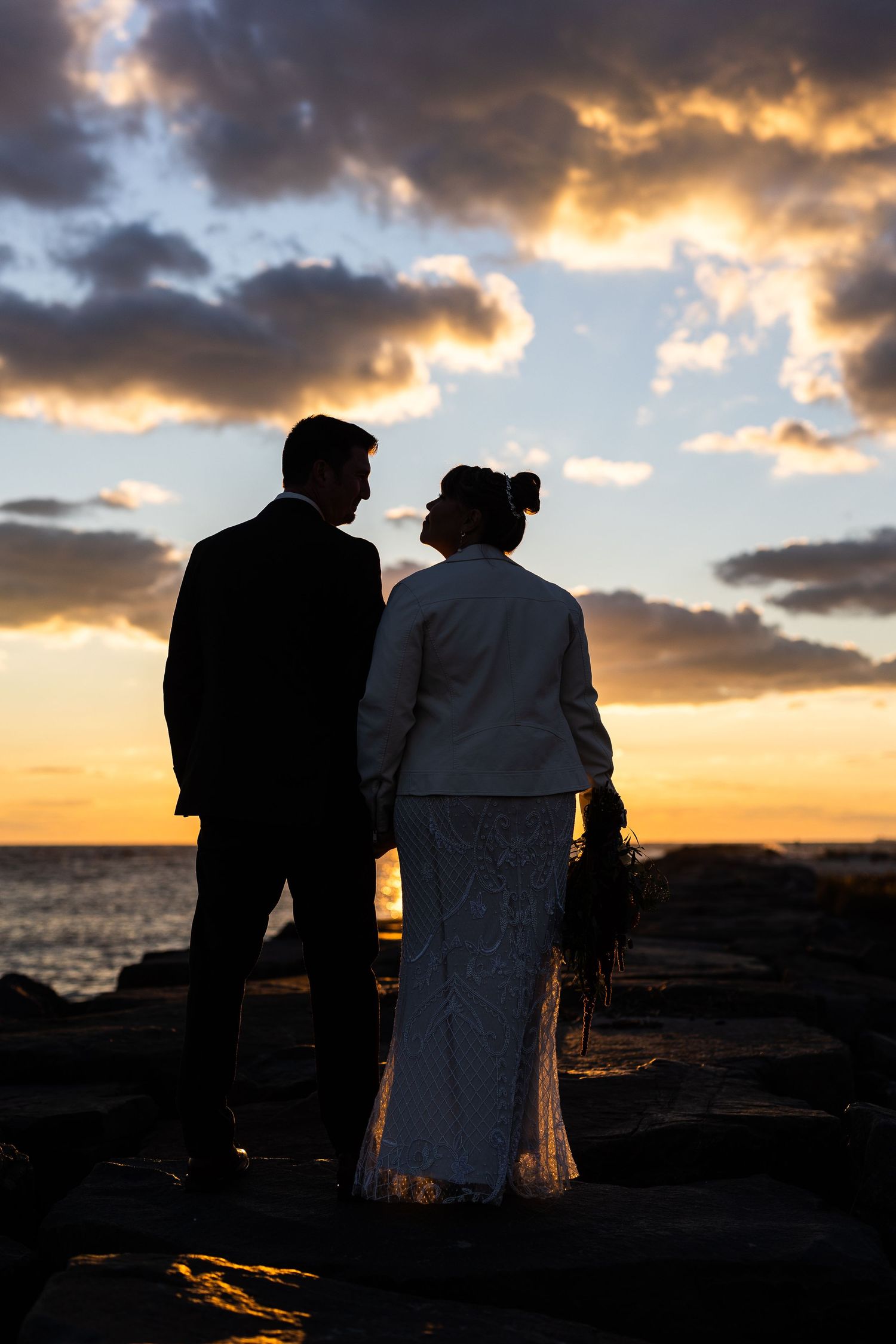 Silhouette of bride and groom holding hands on rocky shoreline against dramatic sunset sky.