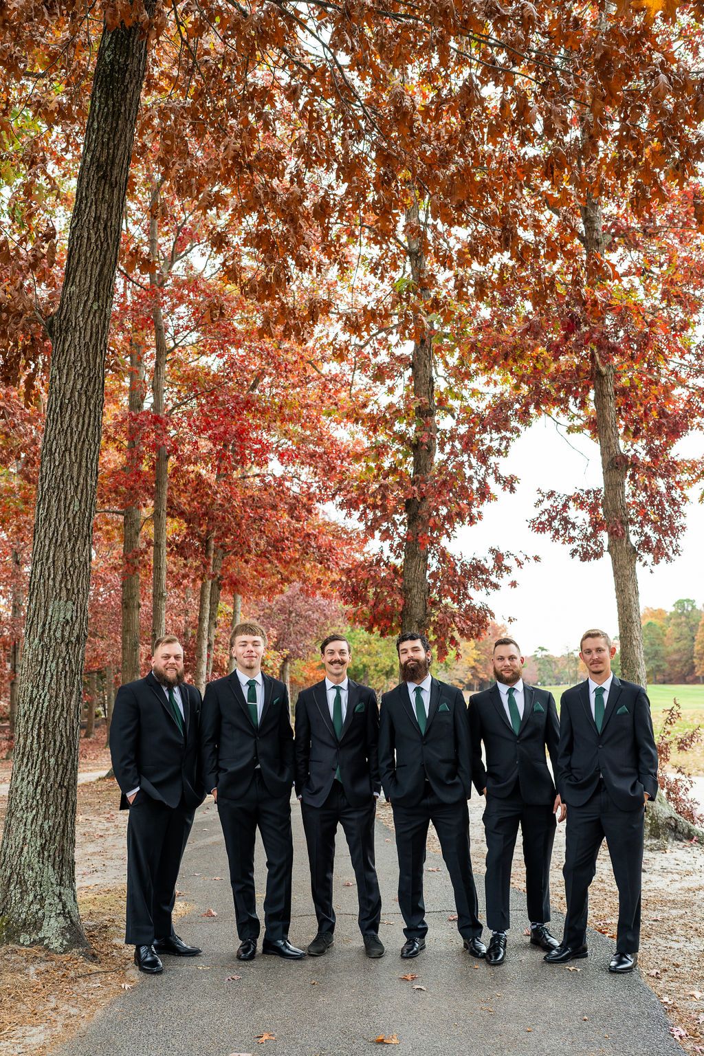 Six groomsmen in black suits with green ties posing on a path surrounded by fall foliage.