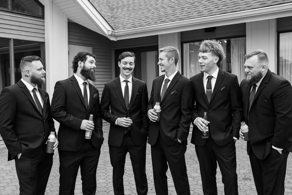 Six groomsmen in black suits laughing together outside a building in black and white wedding photo.