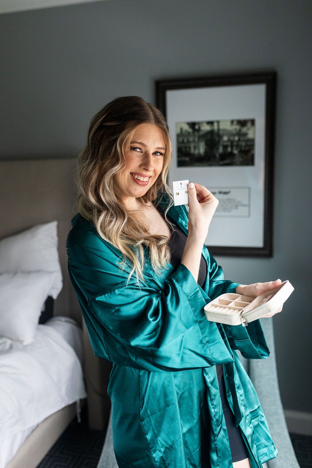 Smiling bride in a teal satin robe holding jewelry while getting ready in a hotel room on her wedding day.
