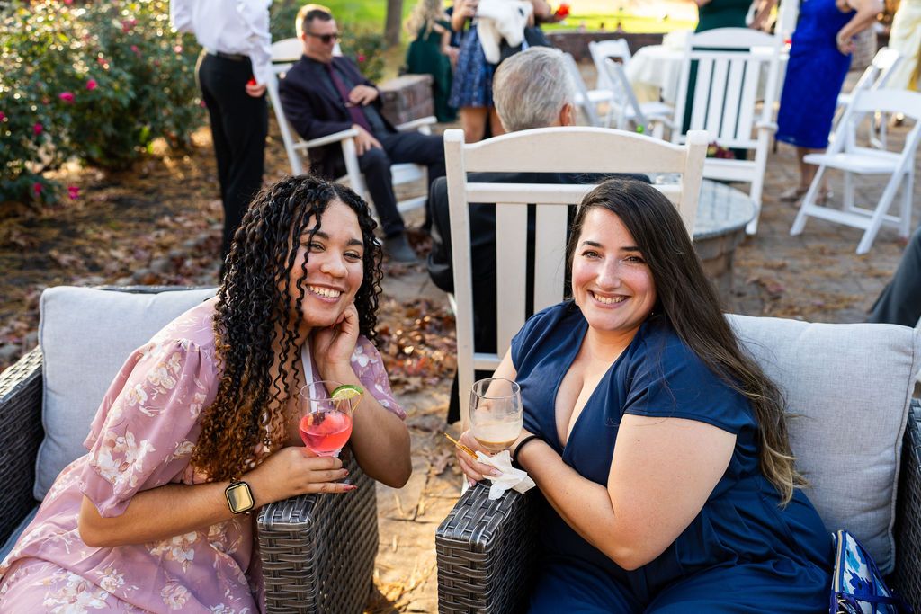Two smiling women holding cocktails seated outdoors at a wedding reception in garden setting.