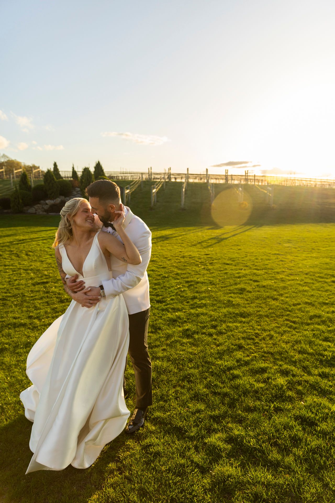 Bride and groom embrace on green lawn with bridge and golden sunset in background.