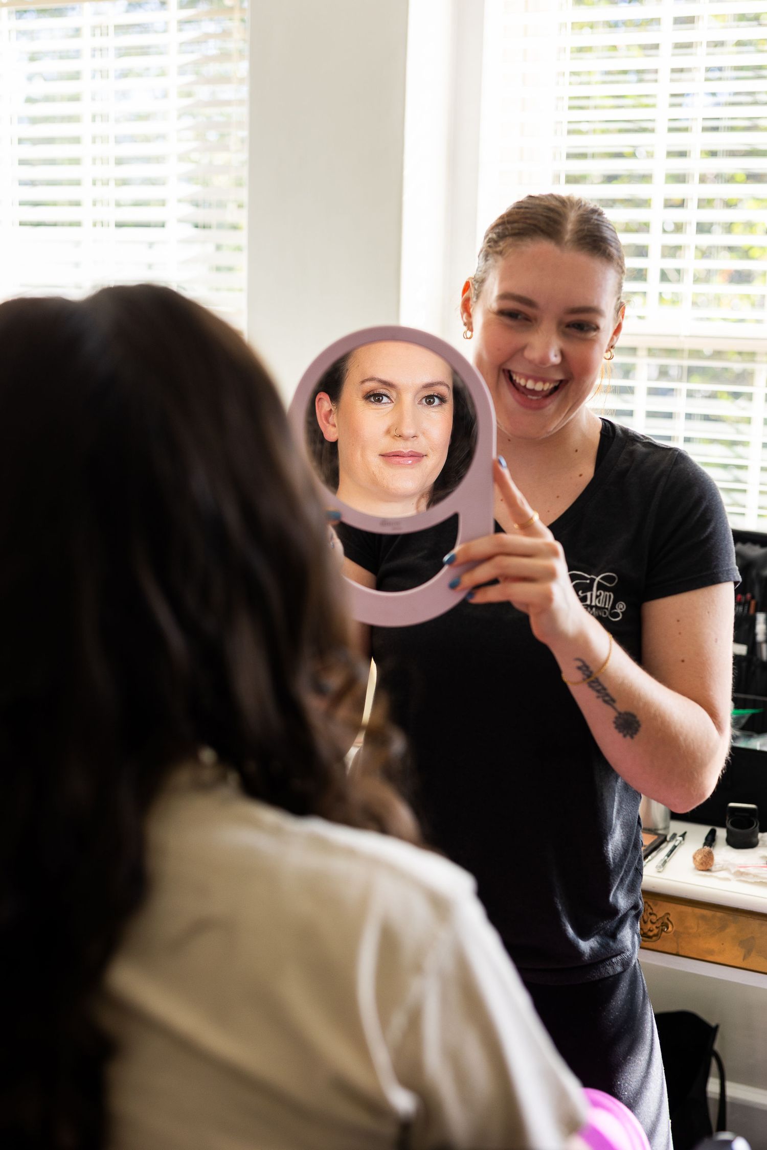 Smiling makeup artist holding a round mirror showing bride's finished wedding makeup look in a bright room.
