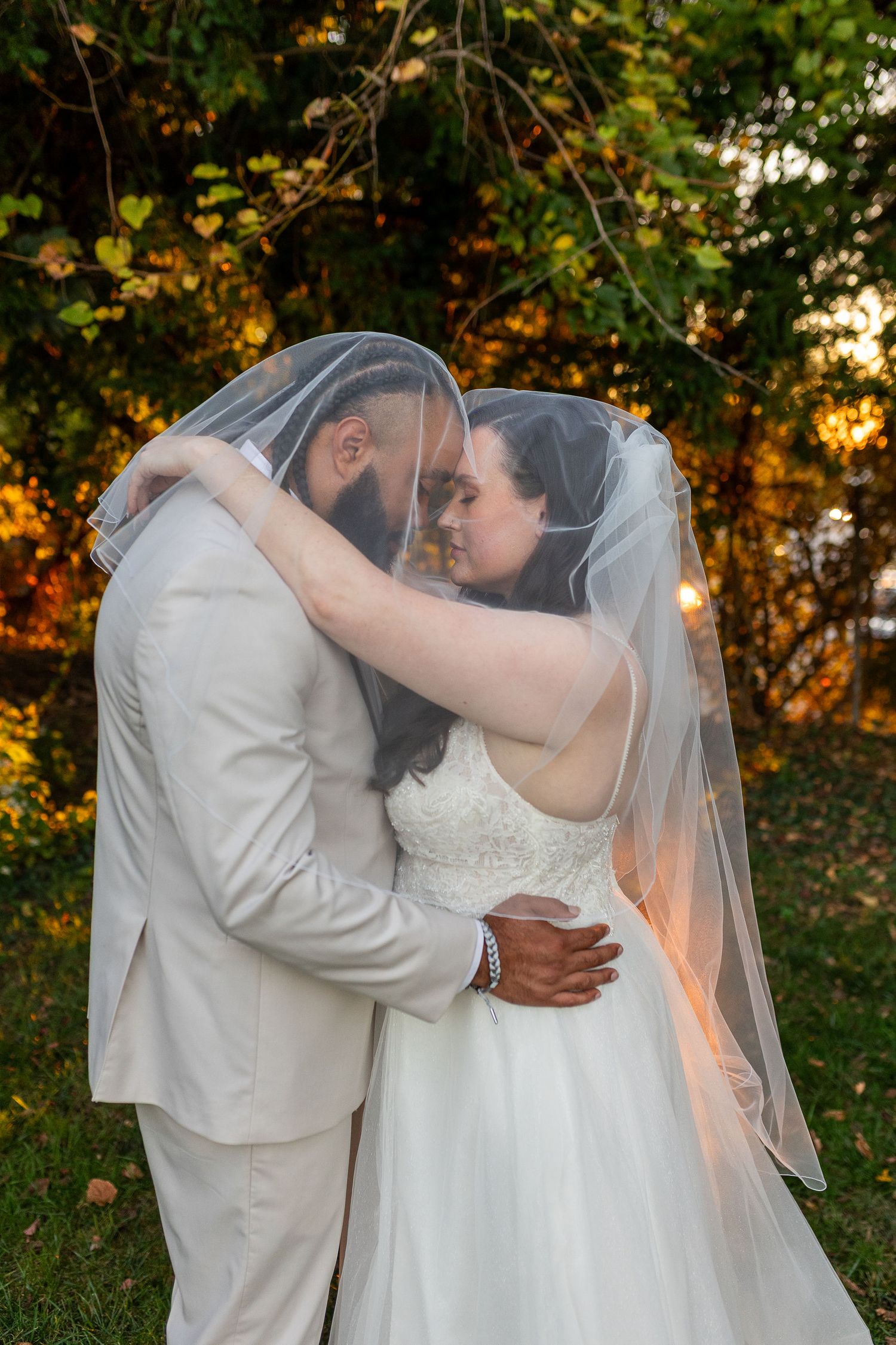 Bride and groom share romantic moment under wedding veil in golden sunset garden setting.