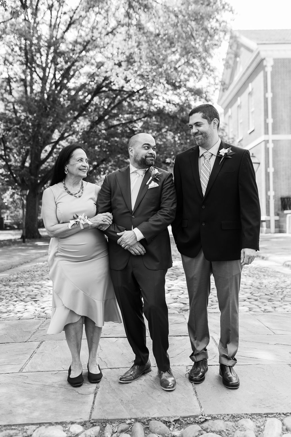 Groom walks with mother and brother in black and white wedding portrait outdoors.