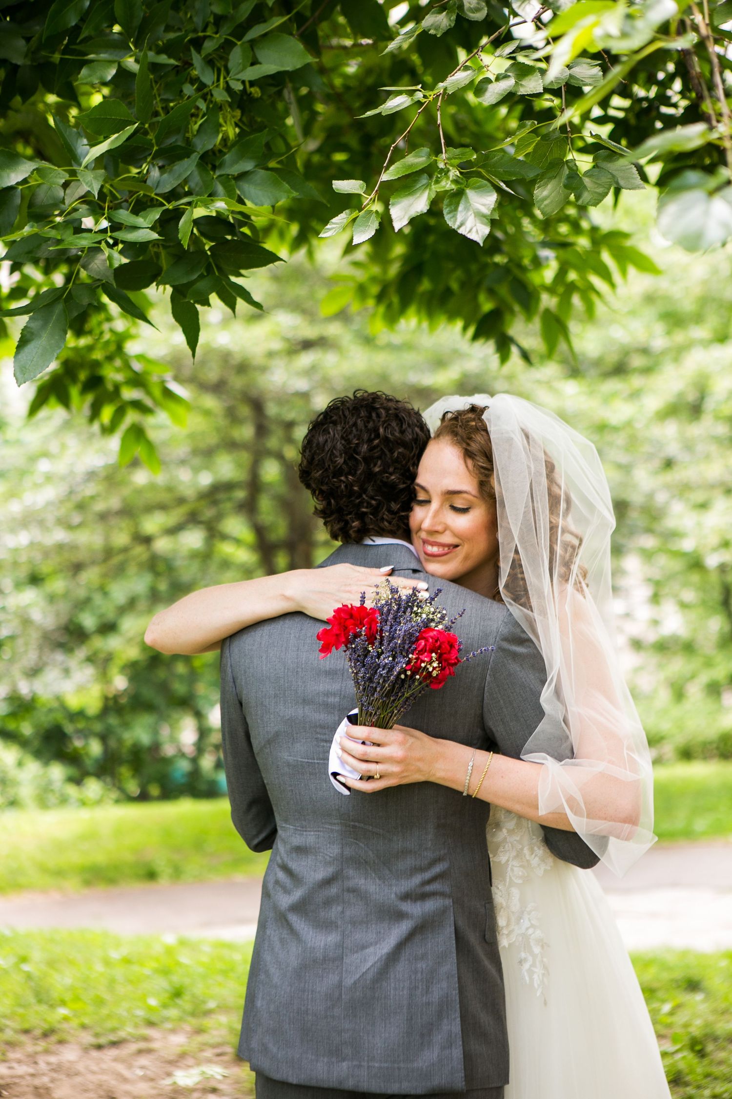Happy bride wearing veil embraces groom in gray suit, clutching red carnation bouquet in lush garden setting.
