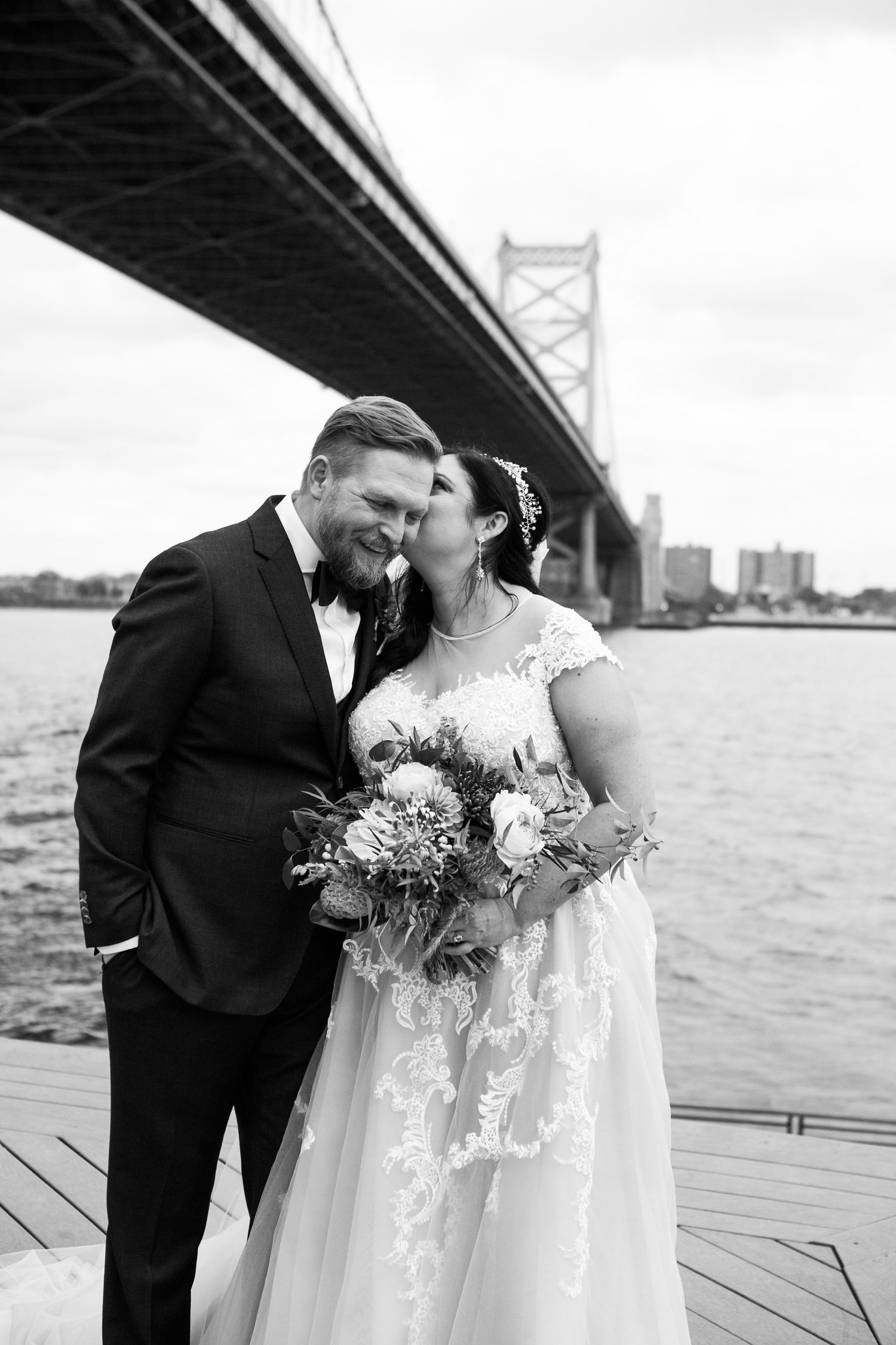 Black and white photo of bride kissing groom on cheek by the Benjamin Franklin Bridge waterfront.