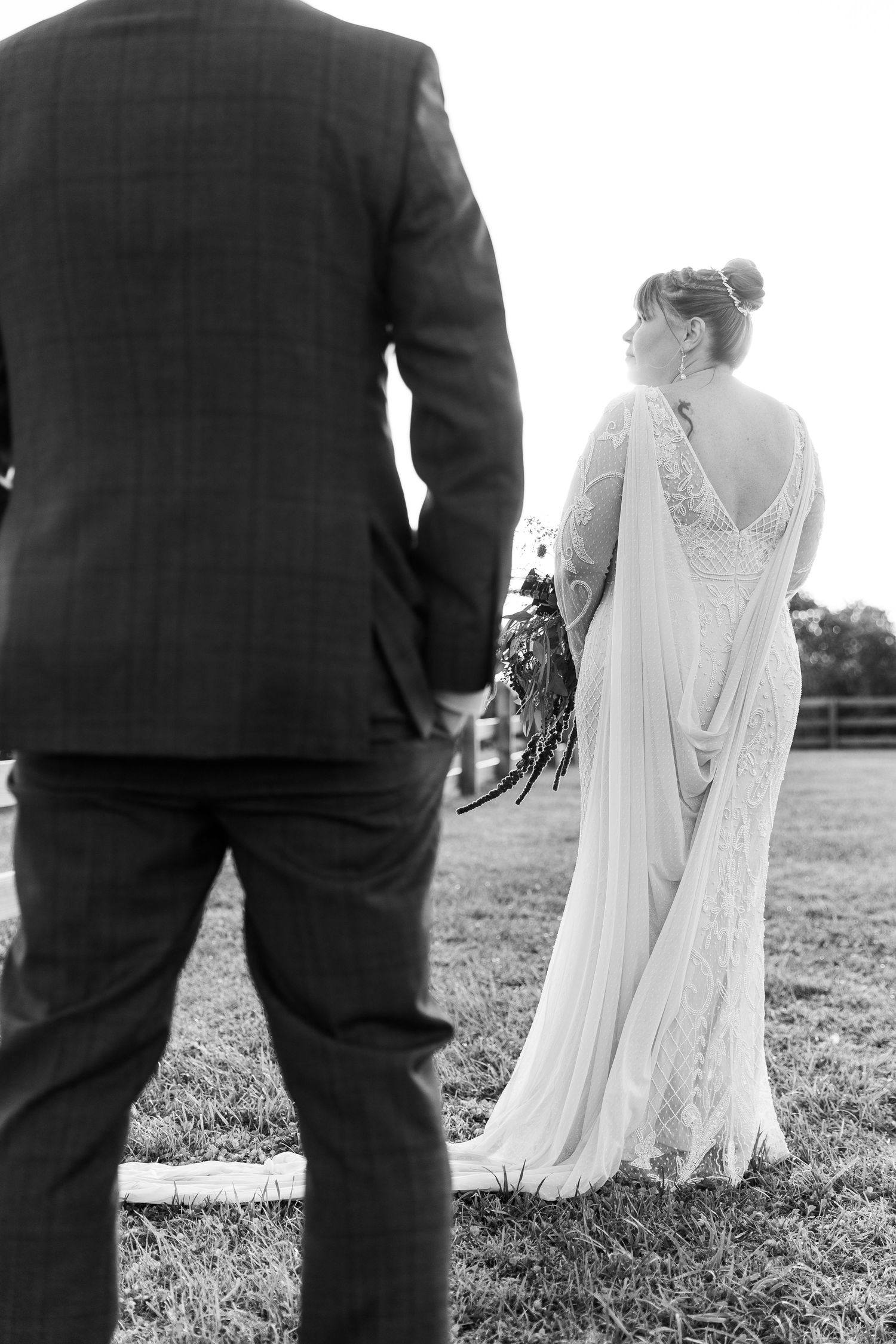 Black and white photo of groom in plaid suit watching bride in lace gown with bouquet outdoors.