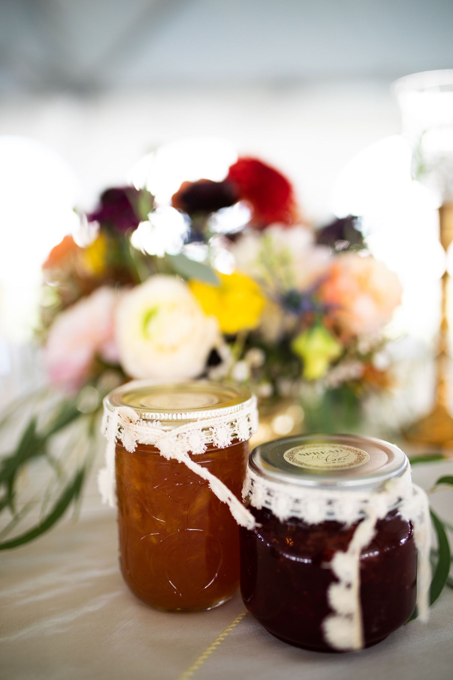 Two rustic mason jars of homemade jam decorated with lace ribbon sit on a white table with colorful floral centerpiece.