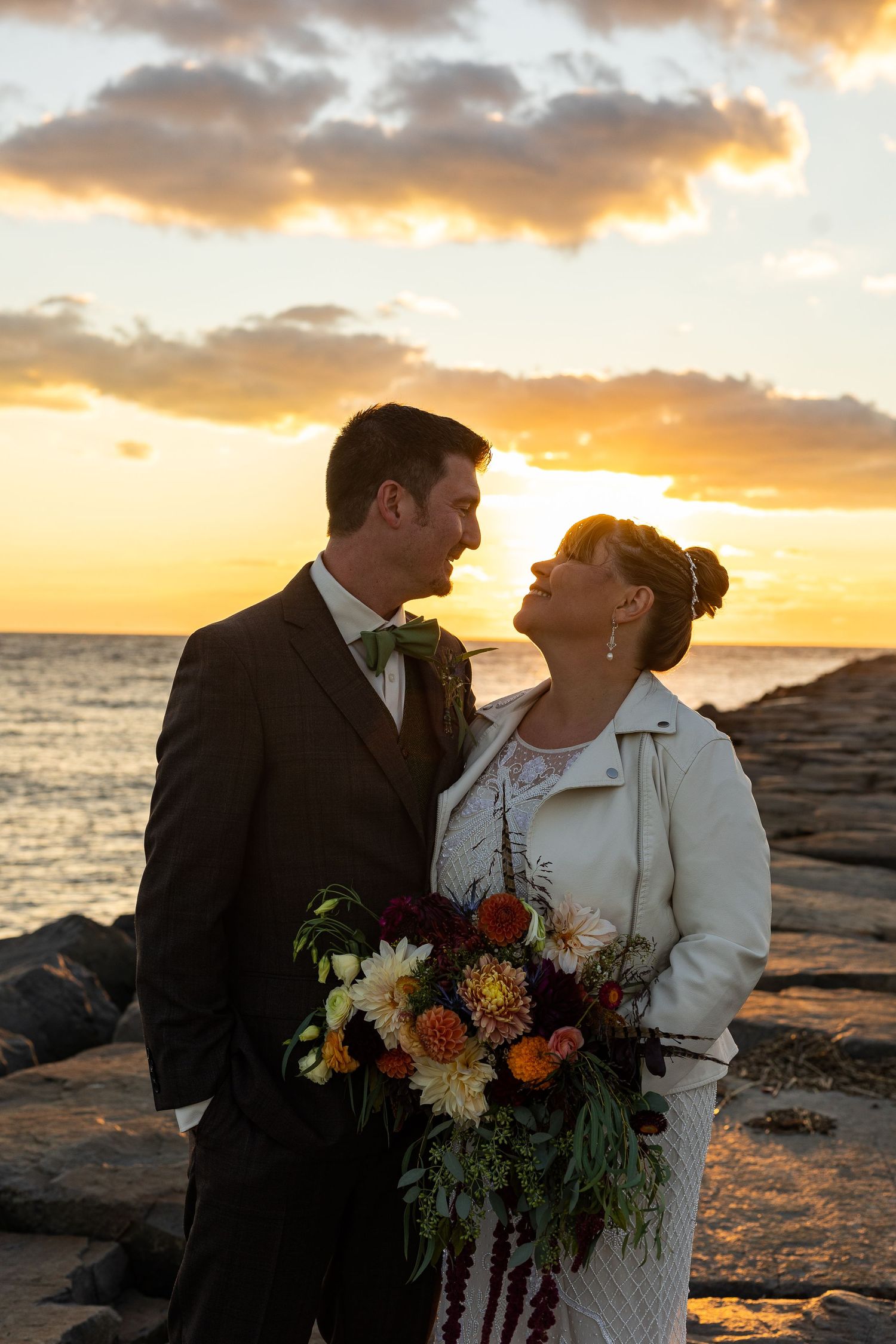 Bride and groom share a romantic moment by the rocky shore during a stunning golden sunset.