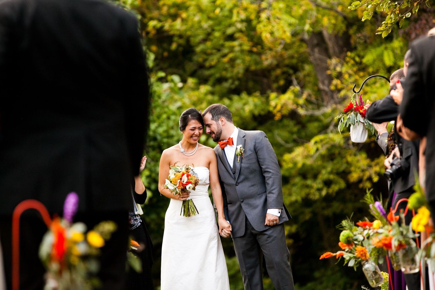 Newlyweds walk down the aisle smiling, surrounded by colorful floral arrangements at outdoor ceremony.