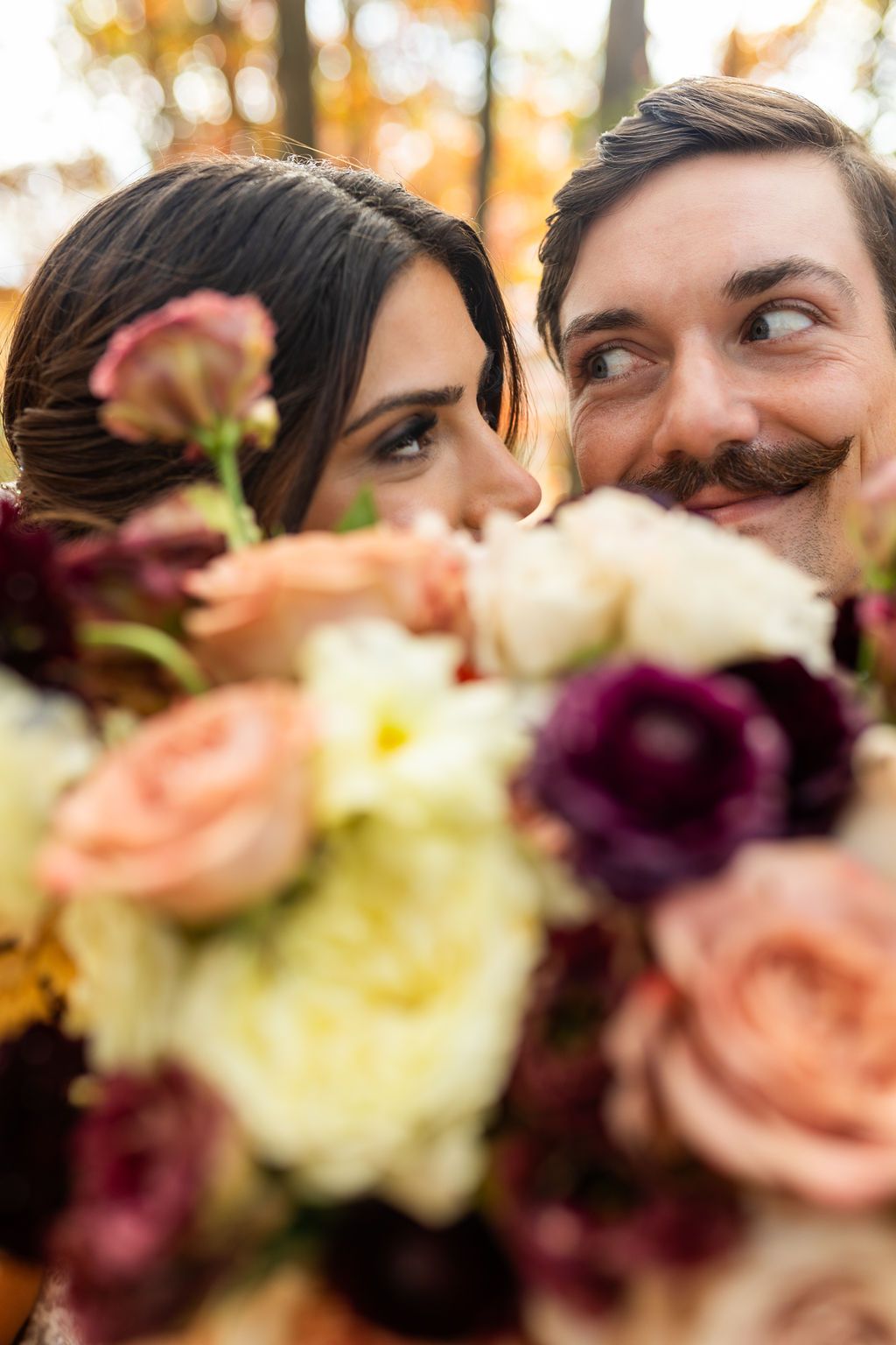 Bride and mustachioed groom peek playfully over a colorful bouquet of roses and dahlias.