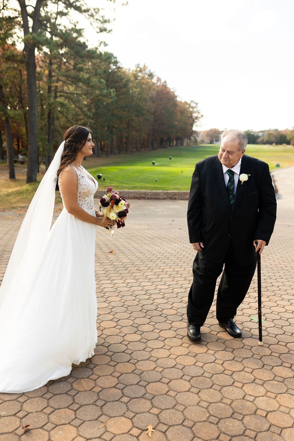 Bride in lace gown meeting elderly gentleman with cane on a brick pathway at outdoor venue.