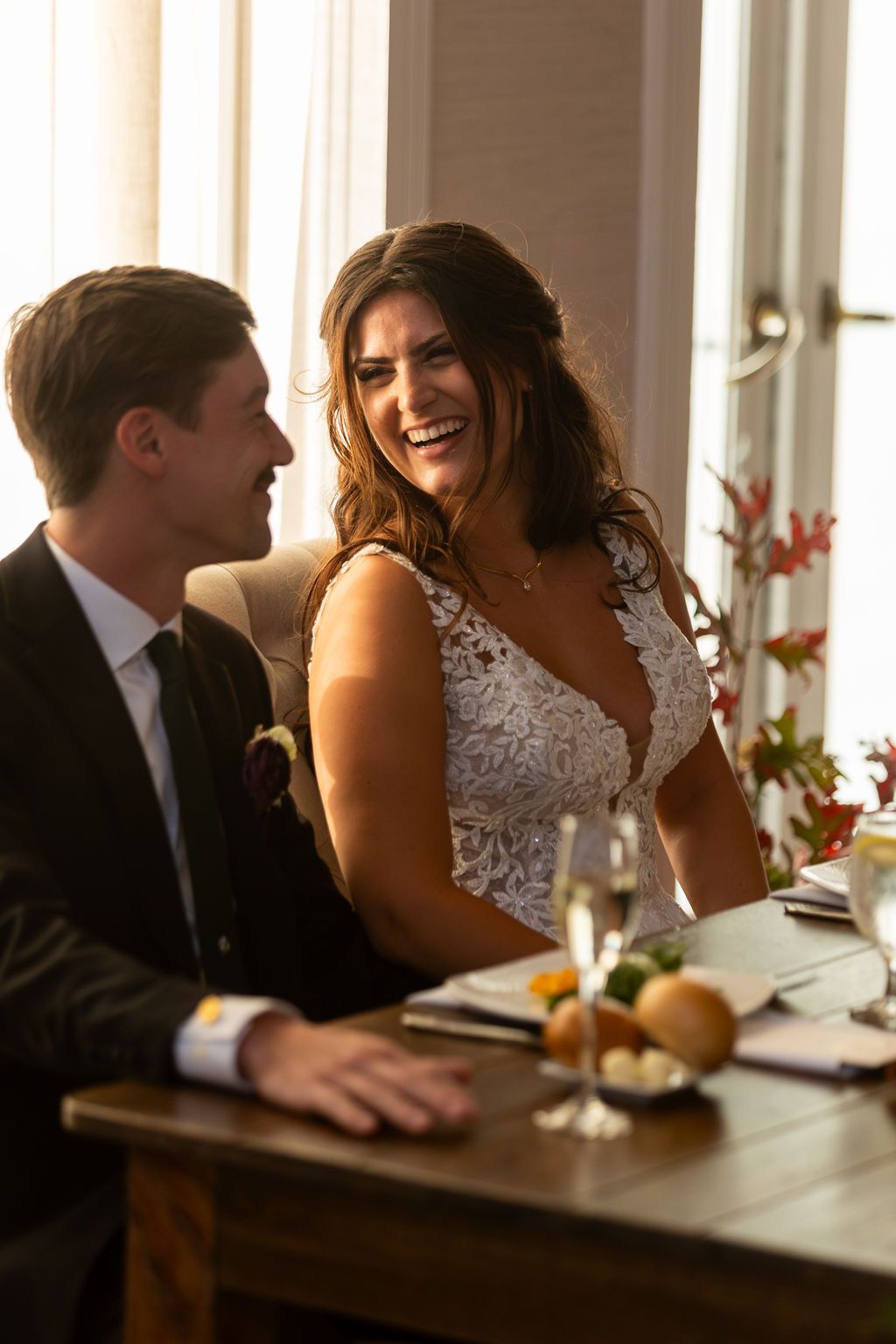 Bride in lace wedding dress laughing joyfully with groom at sweetheart table during wedding reception.