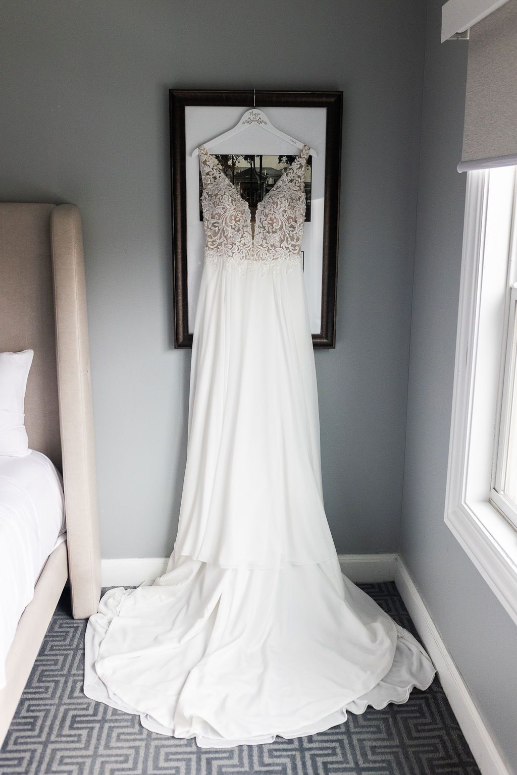 Elegant white wedding gown with lace bodice and flowing skirt hanging on a frame in a hotel room.