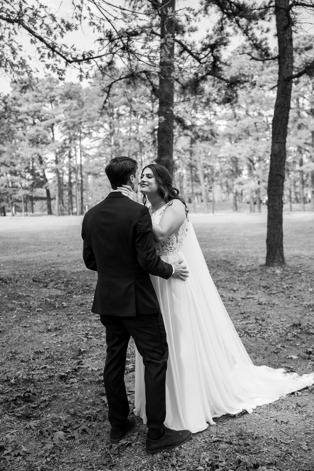 Black and white portrait of bride and groom embracing tenderly beneath pine trees on their wedding day.