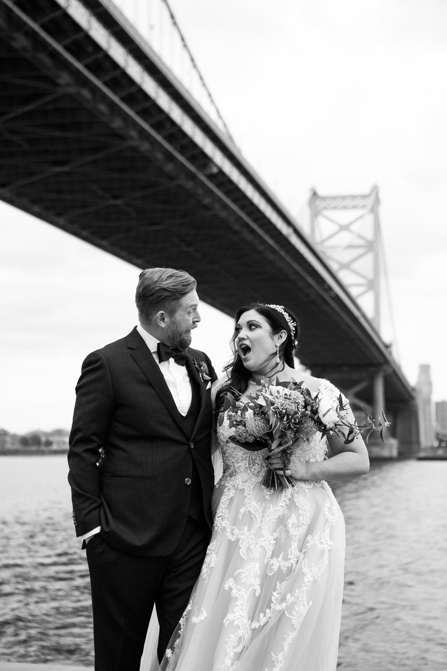Black and white image of laughing bride and groom sharing a playful moment under the Benjamin Franklin Bridge.