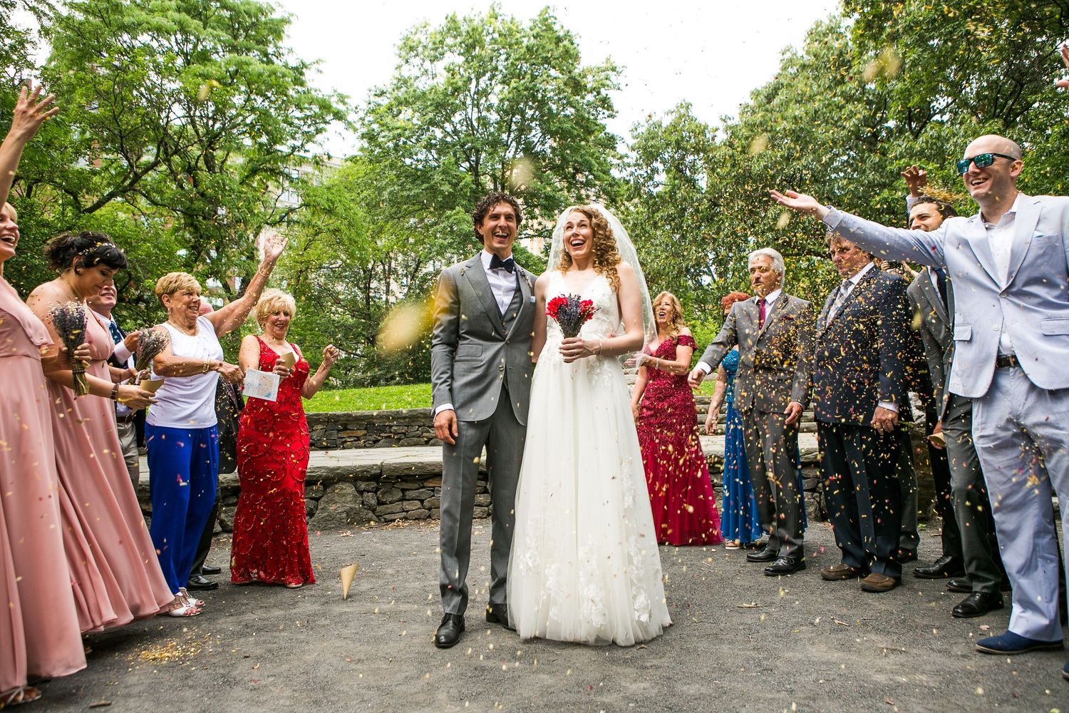 Joyful bride and groom walk through colorful confetti toss surrounded by celebrating wedding guests.