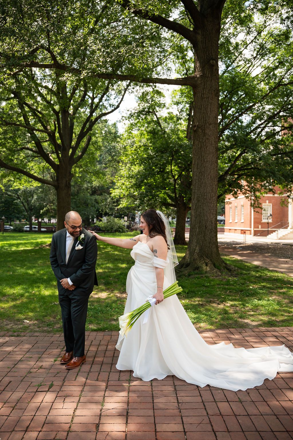 Bride tapping groom on shoulder during romantic first look moment under large trees on brick path.