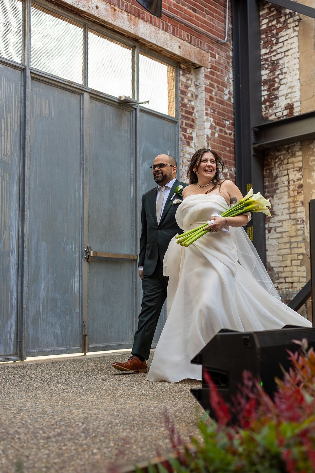 Bride in strapless white gown holding calla lily bouquet walks with groom in dark suit at wedding.